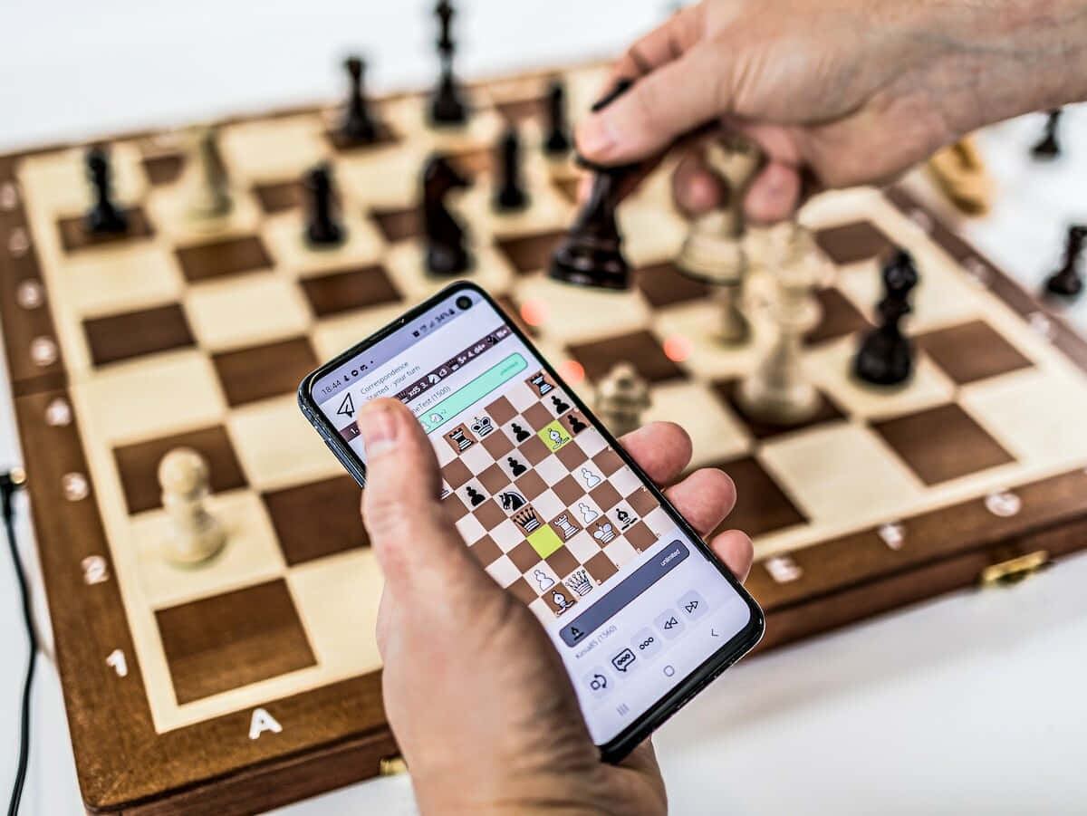 Chessboard 桌布 壁紙 — 一個人下棋，融合實體棋盤和智慧型手機應用程式，專注的手放在木棋盤和發光螢幕上。