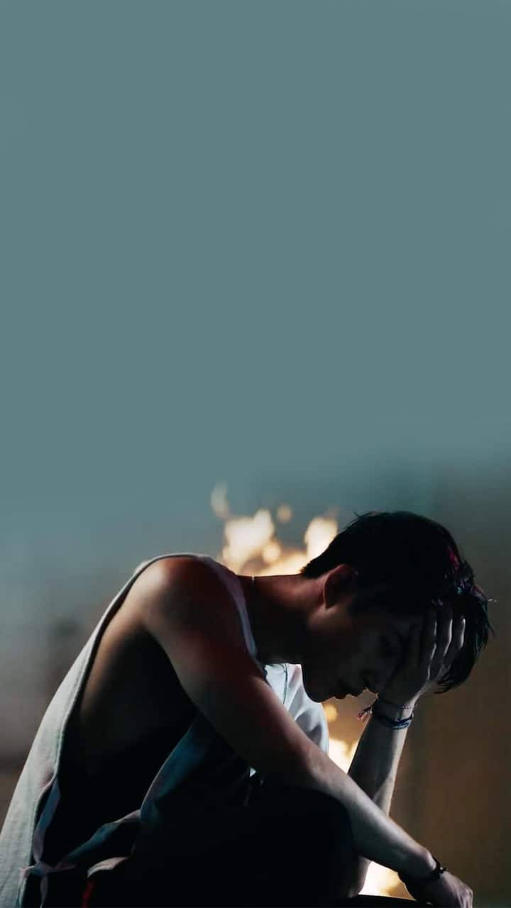 kim hanbin 桌布 壁紙 — 一個人以剪影姿態坐著，雙手抱頭，蜷縮在微弱的青綠色天空下，背景是發光的火焰。