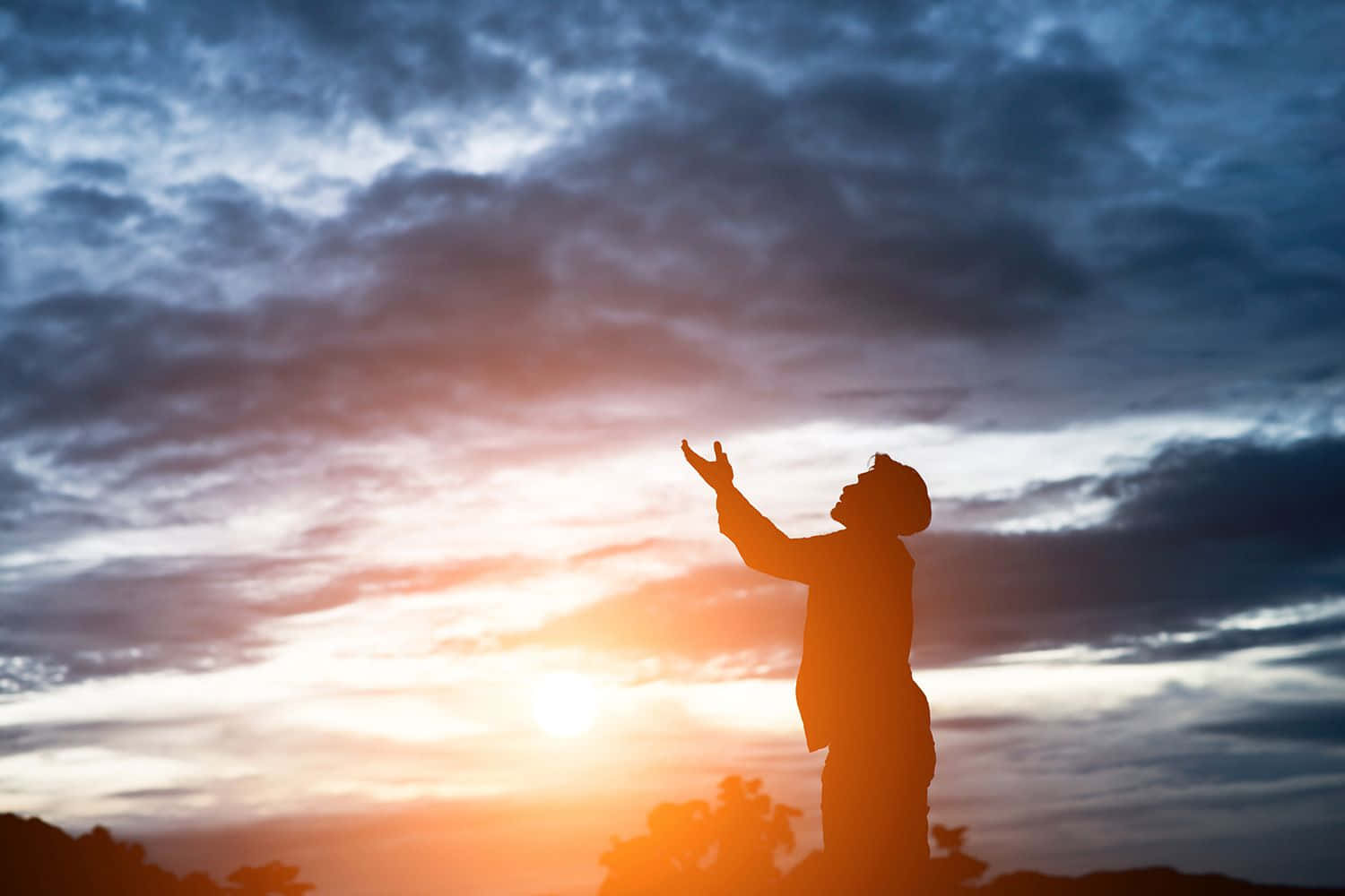 Praying 桌布 壁紙 — 一個人以剪影姿態站立，雙臂高舉向著以火橙色和深藍色雲彩繪成的戲劇性日落天空。