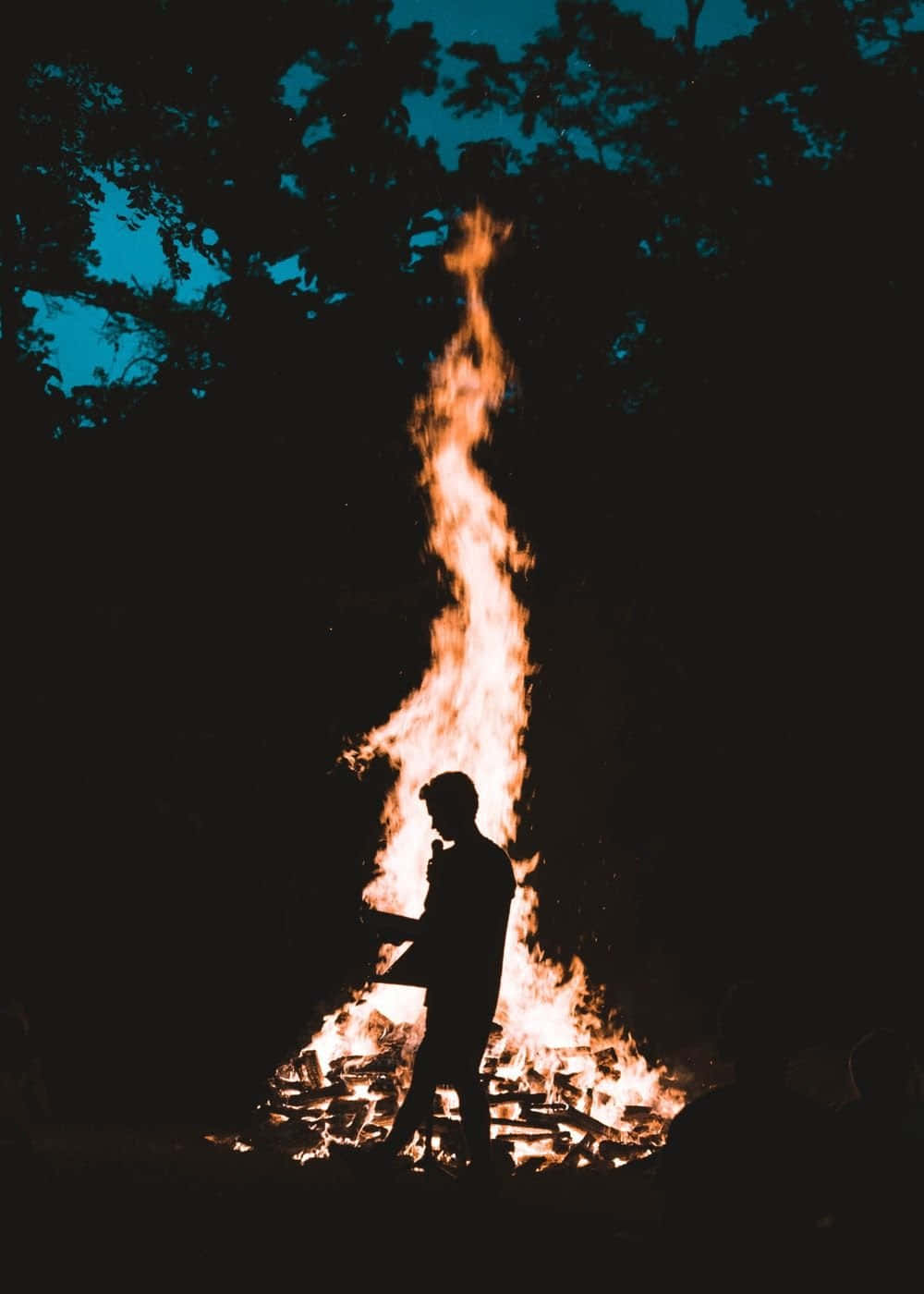 Silhouette 桌布 壁紙 — 一個人以剪影站在一個高聳的營火旁，營火在暮色中對著黑暗的森林發光。