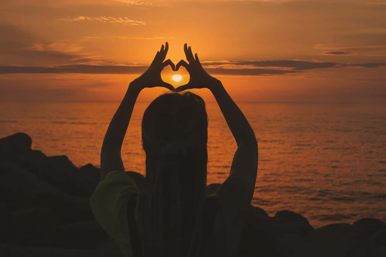 Heart Silhouette 桌布 壁紙 — 一個人以火熱的日落為背景，雙手圍繞著海洋上發光的太陽形成心形。
