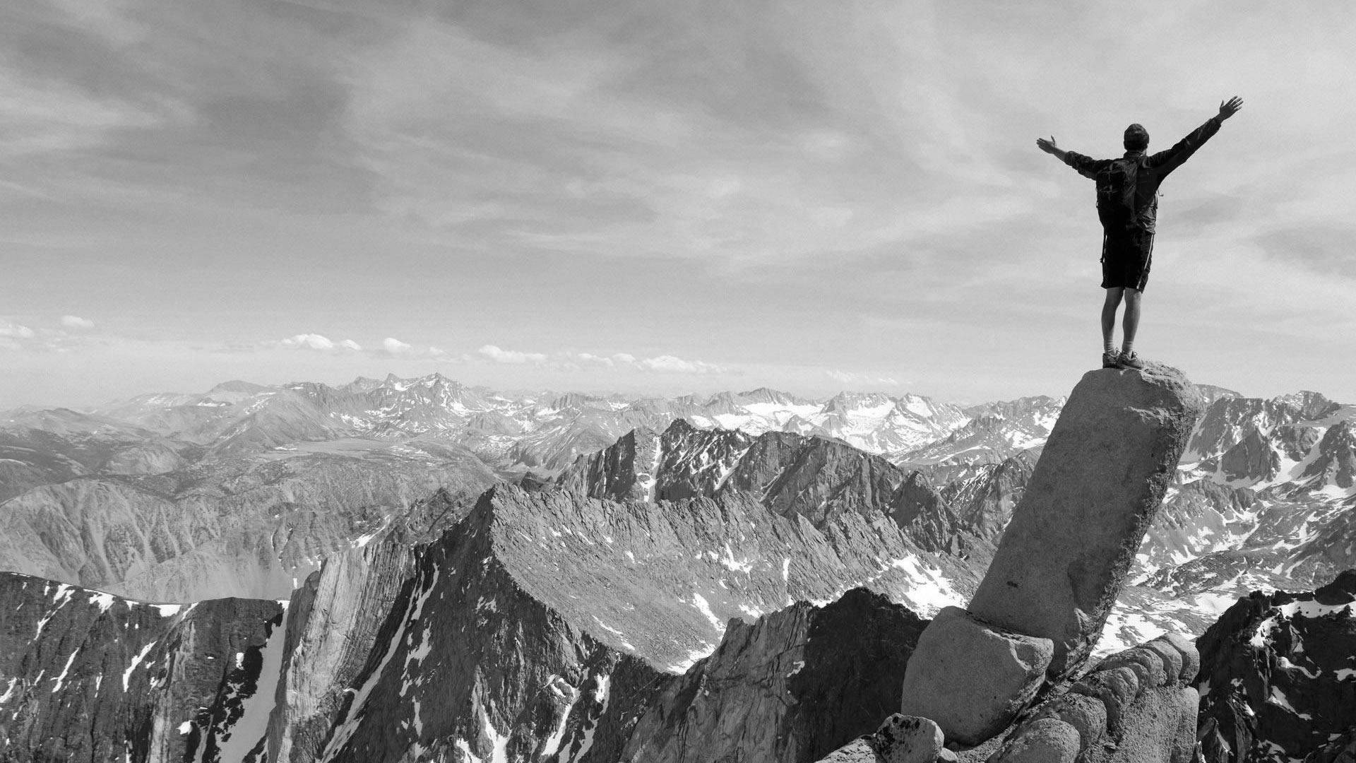 Climbing 桌布 壁紙 — 一個人勝利地站在岩石山頂上，雙臂伸展，俯瞰黑白相間的廣闊雪山脈。