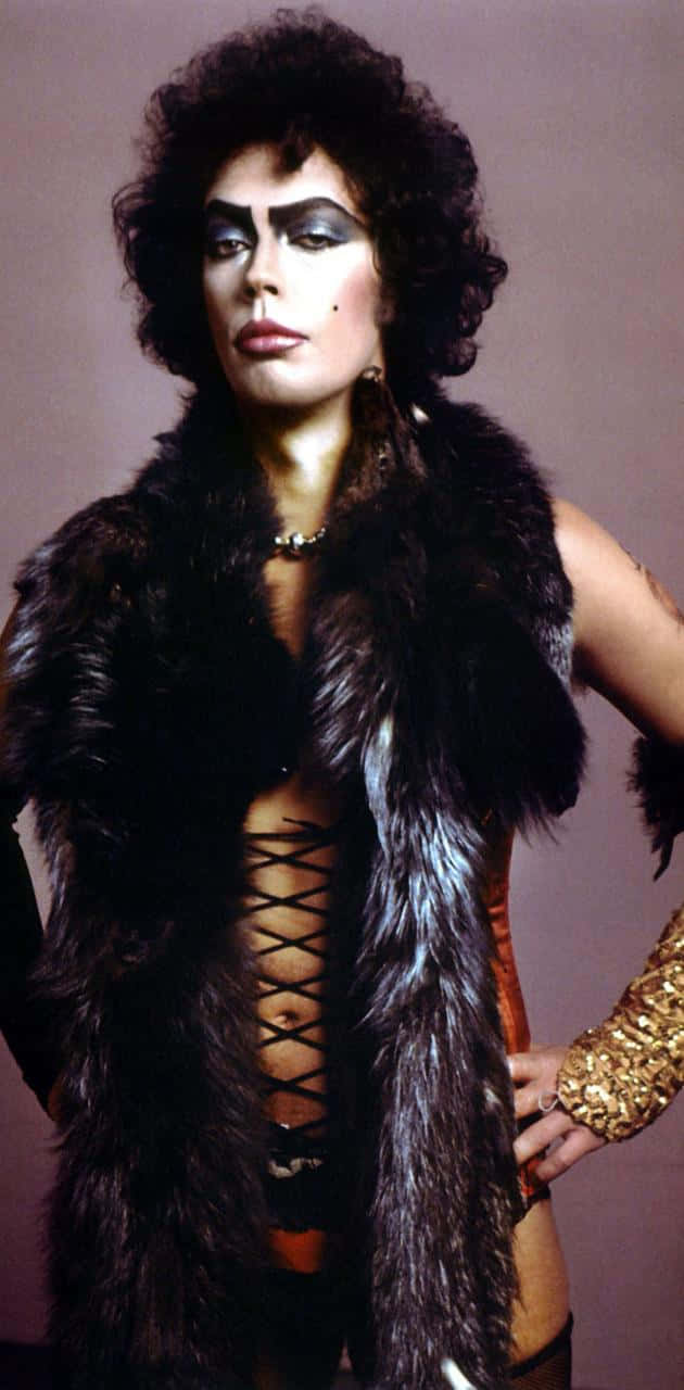Tim Curry 桌布 壁紙 — 一個人化著戲劇性的妝容，擁有豐盈的捲髮，在柔和的背景前，穿著一件奢華的深色毛皮背心，內搭束身衣套裝。