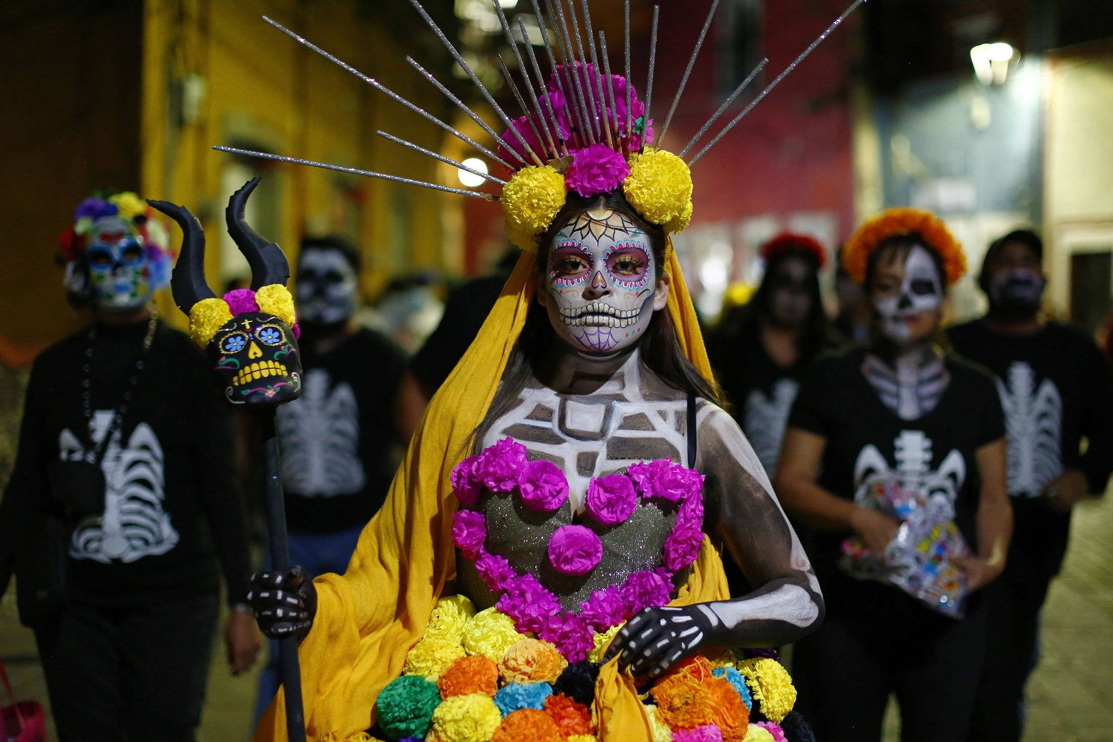 Mexican 桌布 壁紙 — 一個人化著鮮豔的亡靈節妝容，披著黃色披肩，在夜間遊行中手持色彩繽紛的花卉心形。