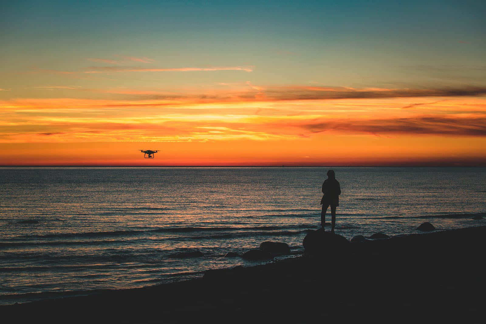 Cute Sunset 桌布 壁紙 — 一個人和一個無人機的剪影，襯著海洋上充滿活力的橙色和藍色日落。