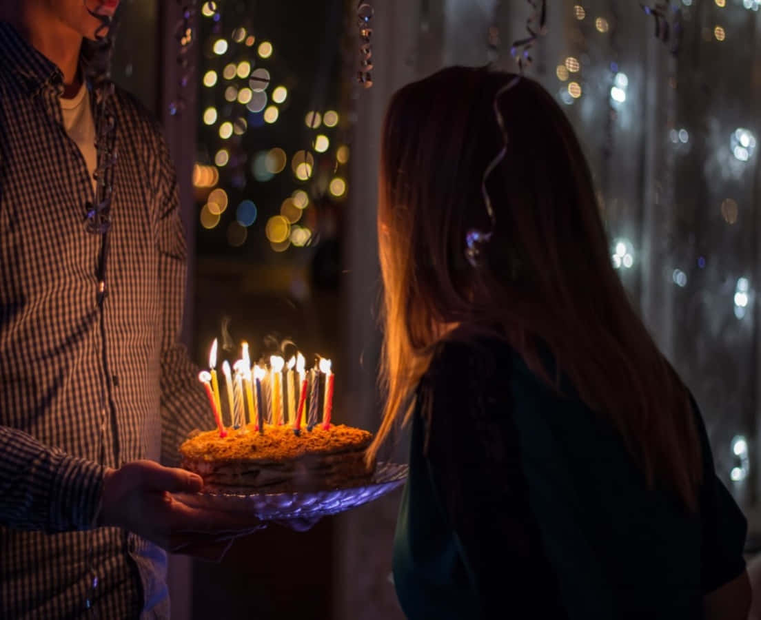 Birthday Celebration 桌布 壁紙 — 一個人在一個溫暖照明的房間裡，在散景燈串下，向另一個人展示一個點燃蠟燭的生日蛋糕。