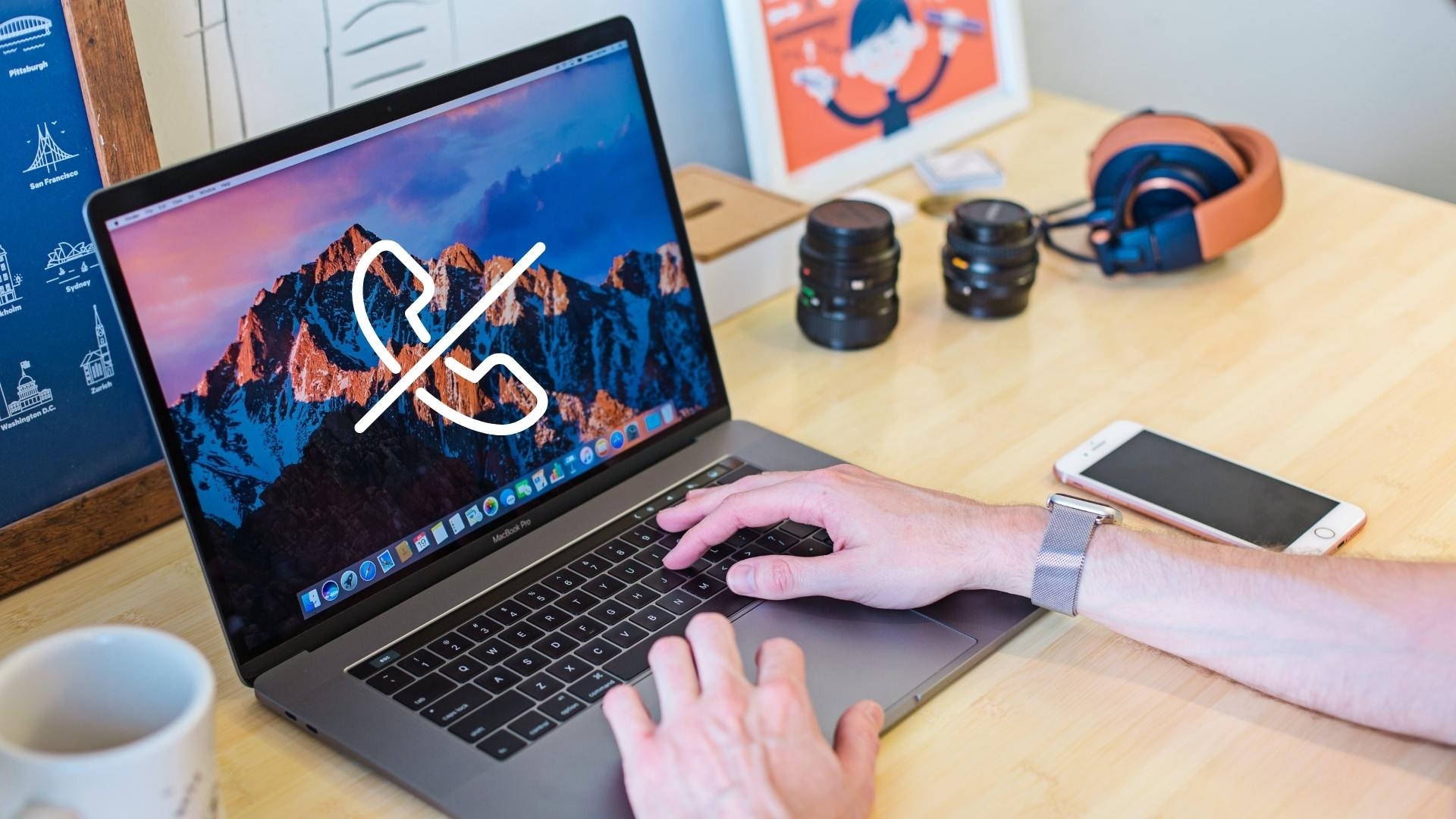 MacBook 動態圖片 桌布 壁紙 — 一個人在一台顯示山脈桌面的筆記型電腦上打字，並在木桌上疊加了一個電話靜音圖標，桌上還有耳機和相機鏡頭。