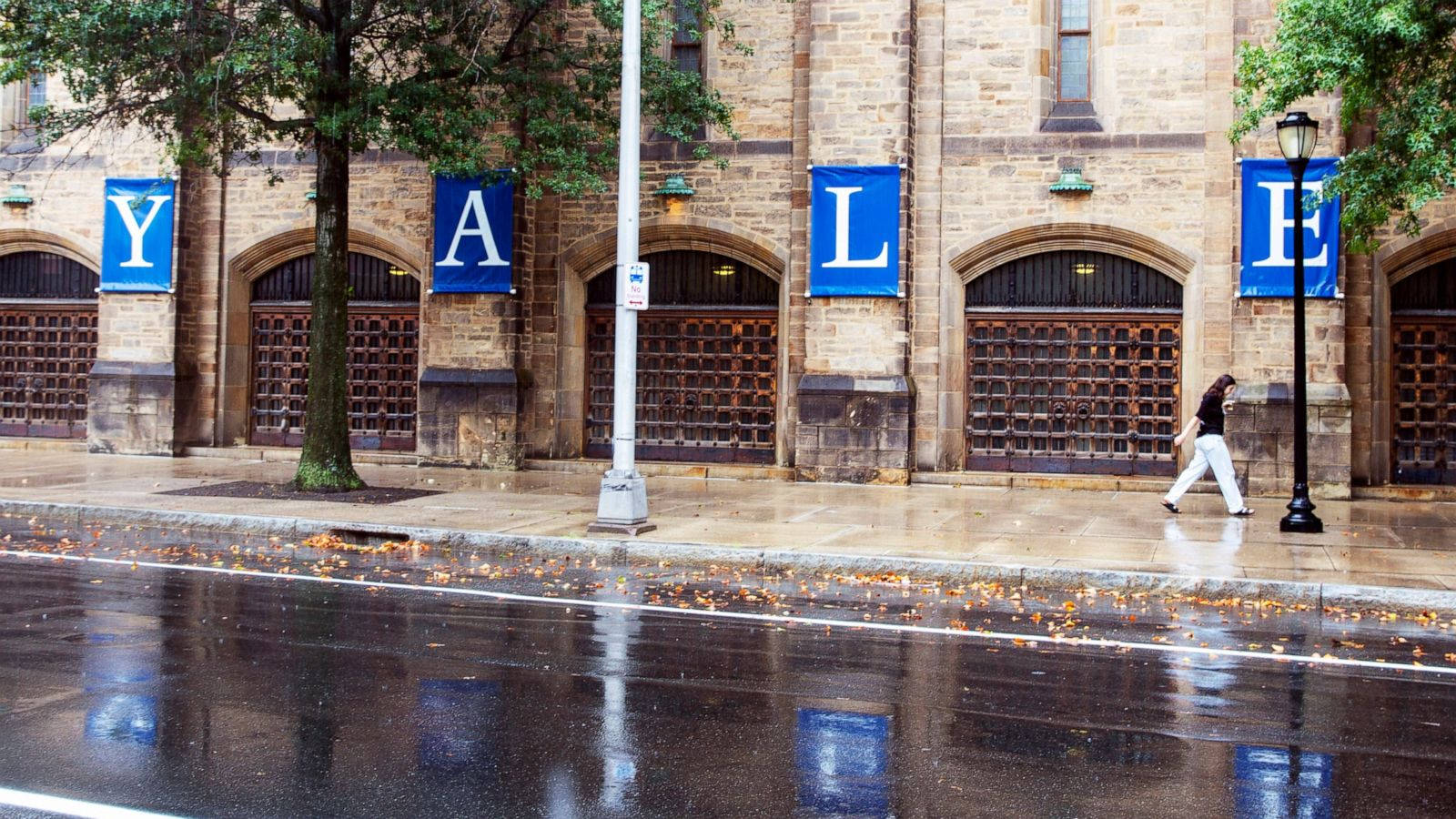 Building 桌布 壁紙 — 一個人在下雨天走過一棟掛有「YALE」橫幅的歷史石建築，濕漉漉的路面倒映著場景。