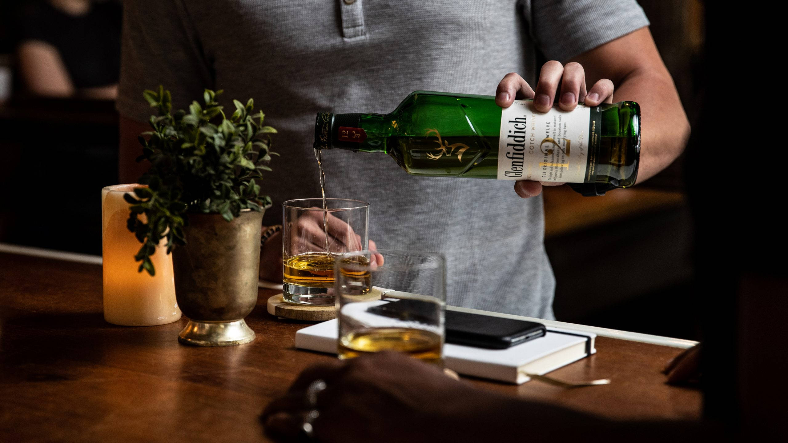 bartender 桌布 壁紙 — 一個人在光線溫暖的木吧檯上，從綠色酒瓶將琥珀色威士忌倒入玻璃杯中，旁邊有蠟燭和筆記本。