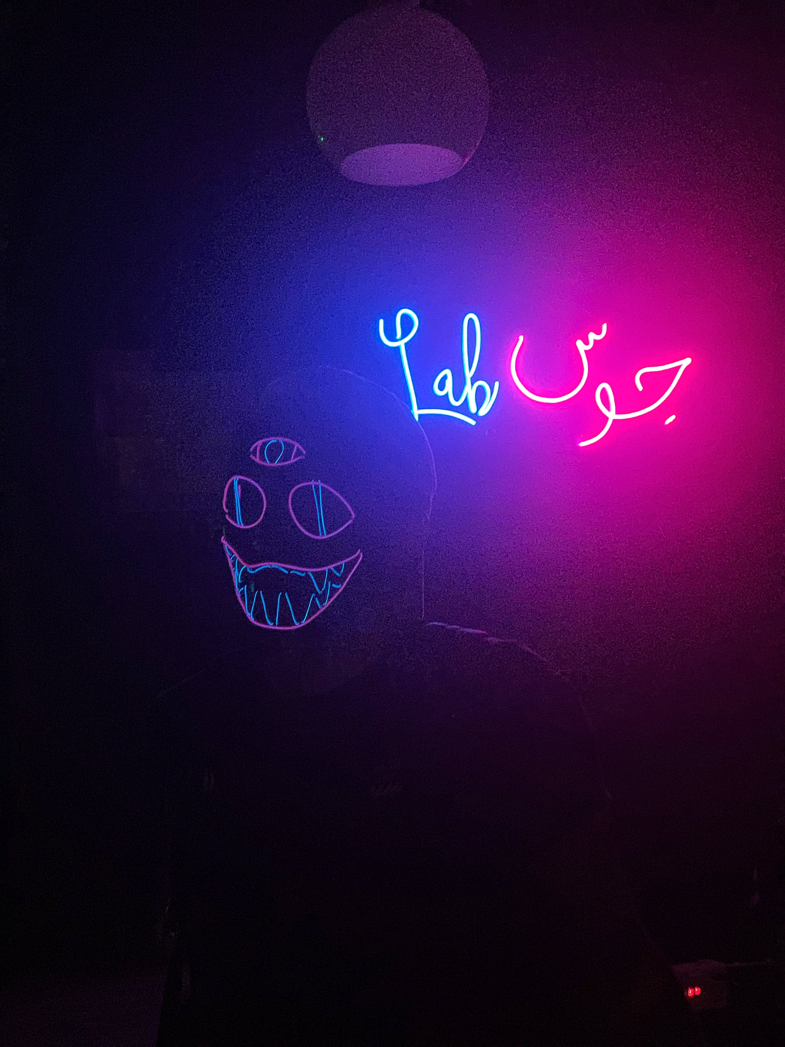 霓虹美學 桌布 壁紙 — 一個人在吊燈下戴著發光的霓虹面具，燈上有 'Lab' 和阿拉伯文字，呈現鮮豔的粉紅色和藍色光。