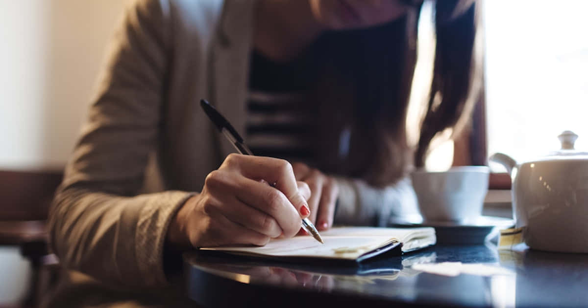 paper 桌布 壁紙 — 一個人在咖啡館的桌子上寫筆記，旁邊放著茶壺和杯子，沐浴在柔和自然光中。