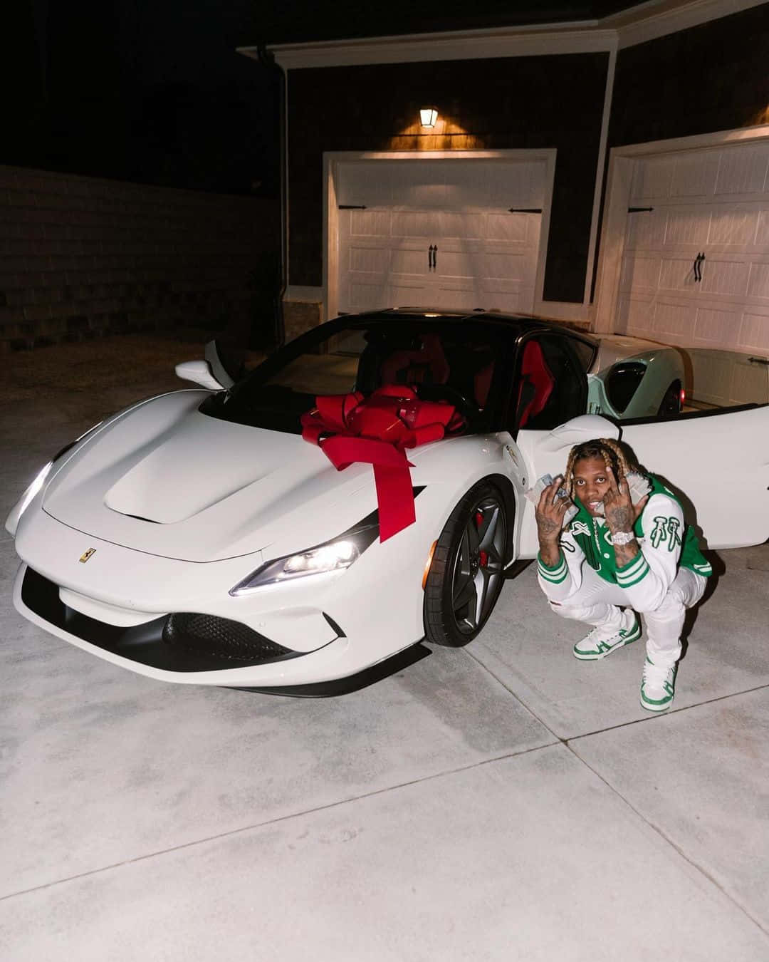 Lil Durk 桌布 壁紙 — 一個人在夜晚燈光通明的車道上蹲在一輛裝飾著巨大紅色蝴蝶結的白色法拉利跑車旁邊。