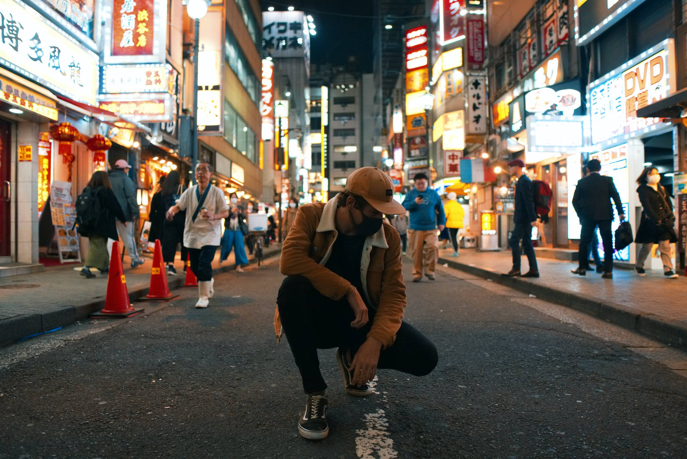 城市 桌布 壁紙 — 一個人在夜晚的東京小巷中蹲下，周圍是發光的招牌和行人。