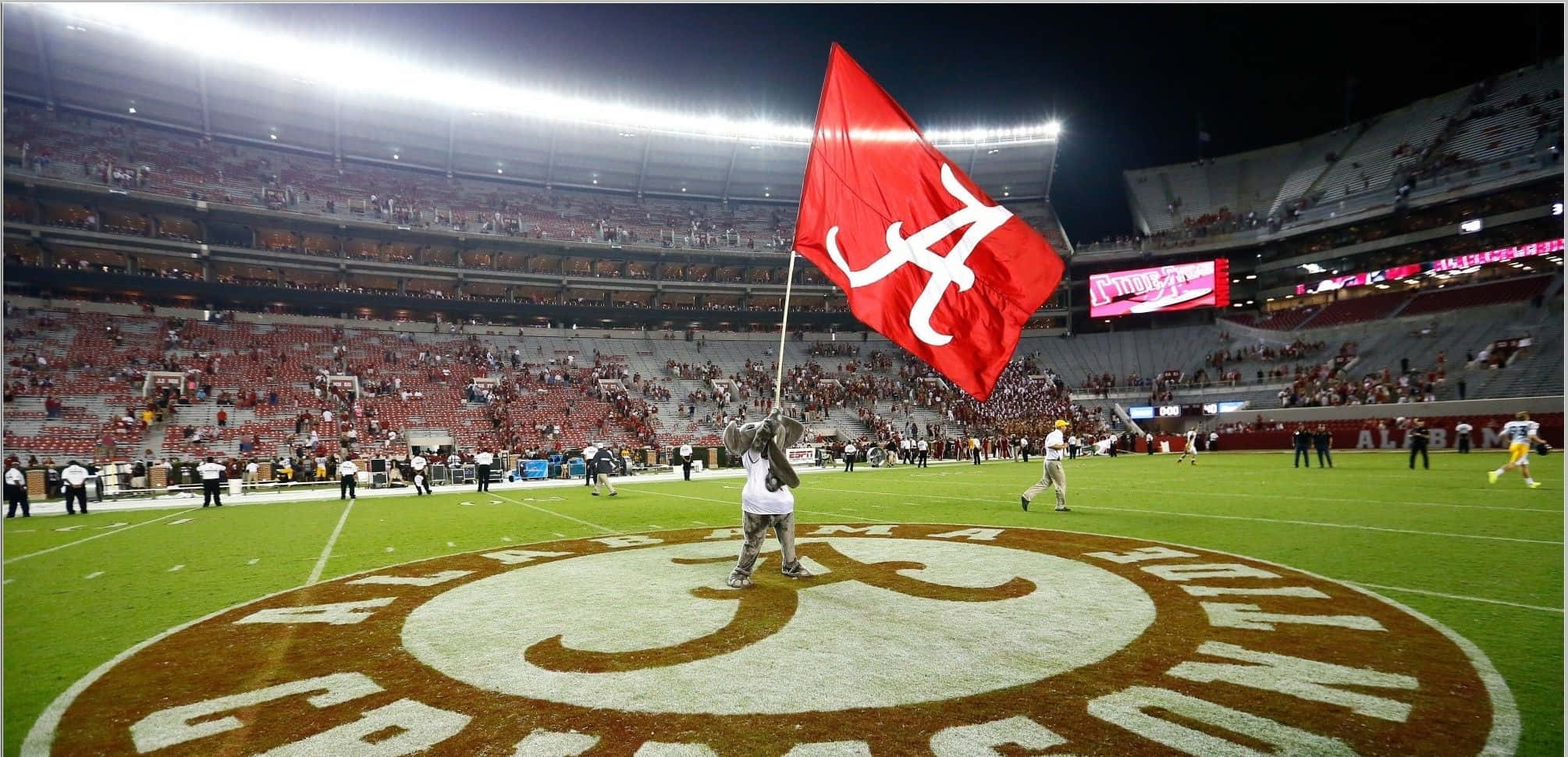 Alabama Football 桌布 壁紙 — 一個人在夜晚的足球場上，在明亮的體育場燈光下，揮舞著深紅色的阿拉巴馬州旗，看台上有球迷。
