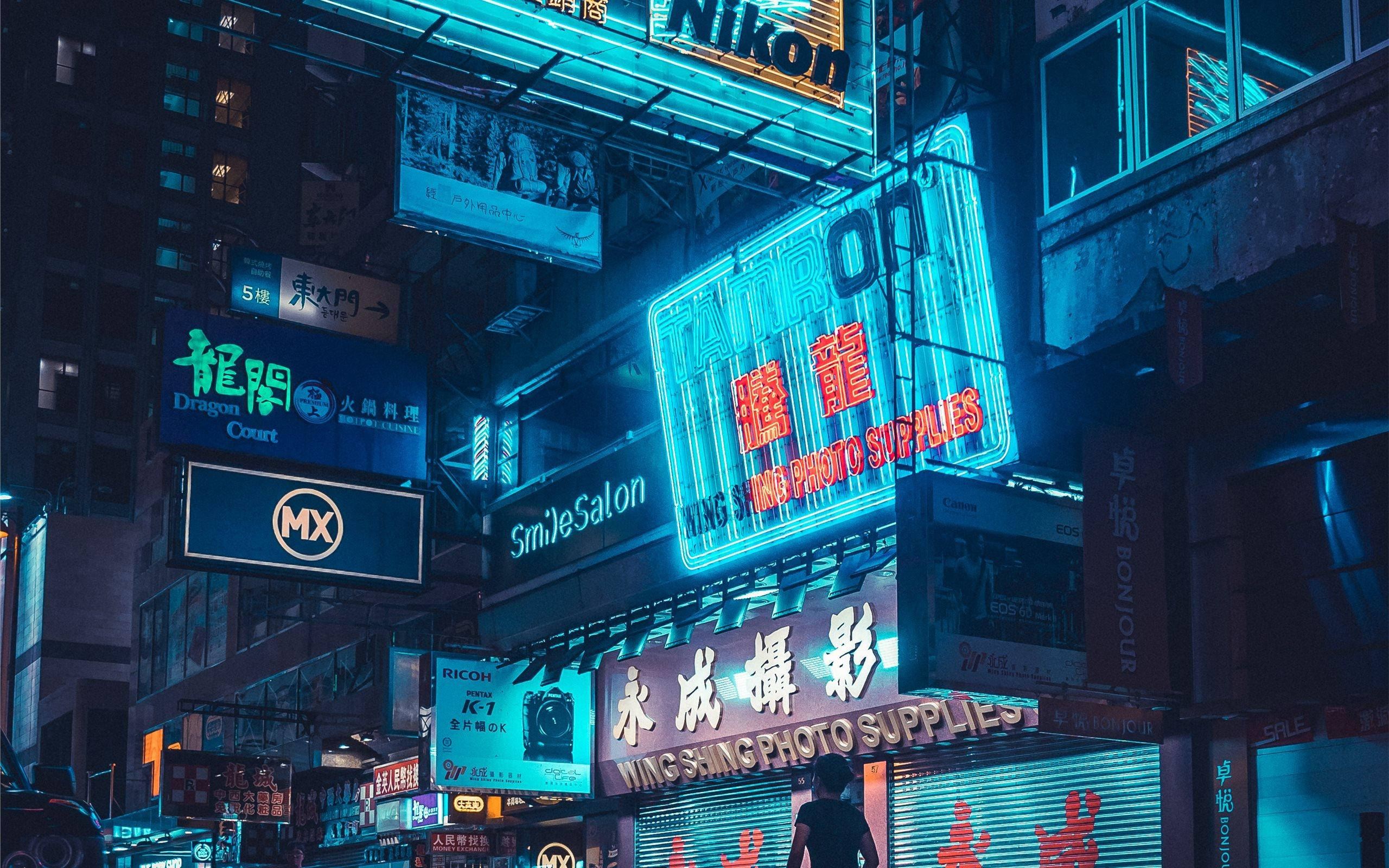 美學 MacBook 壁紙 桌布 壁紙 — 一個人在夜晚走在密集的城市小巷中，頭頂是發光的霓虹燈牌，沐浴在電藍色和紅色的燈光中。