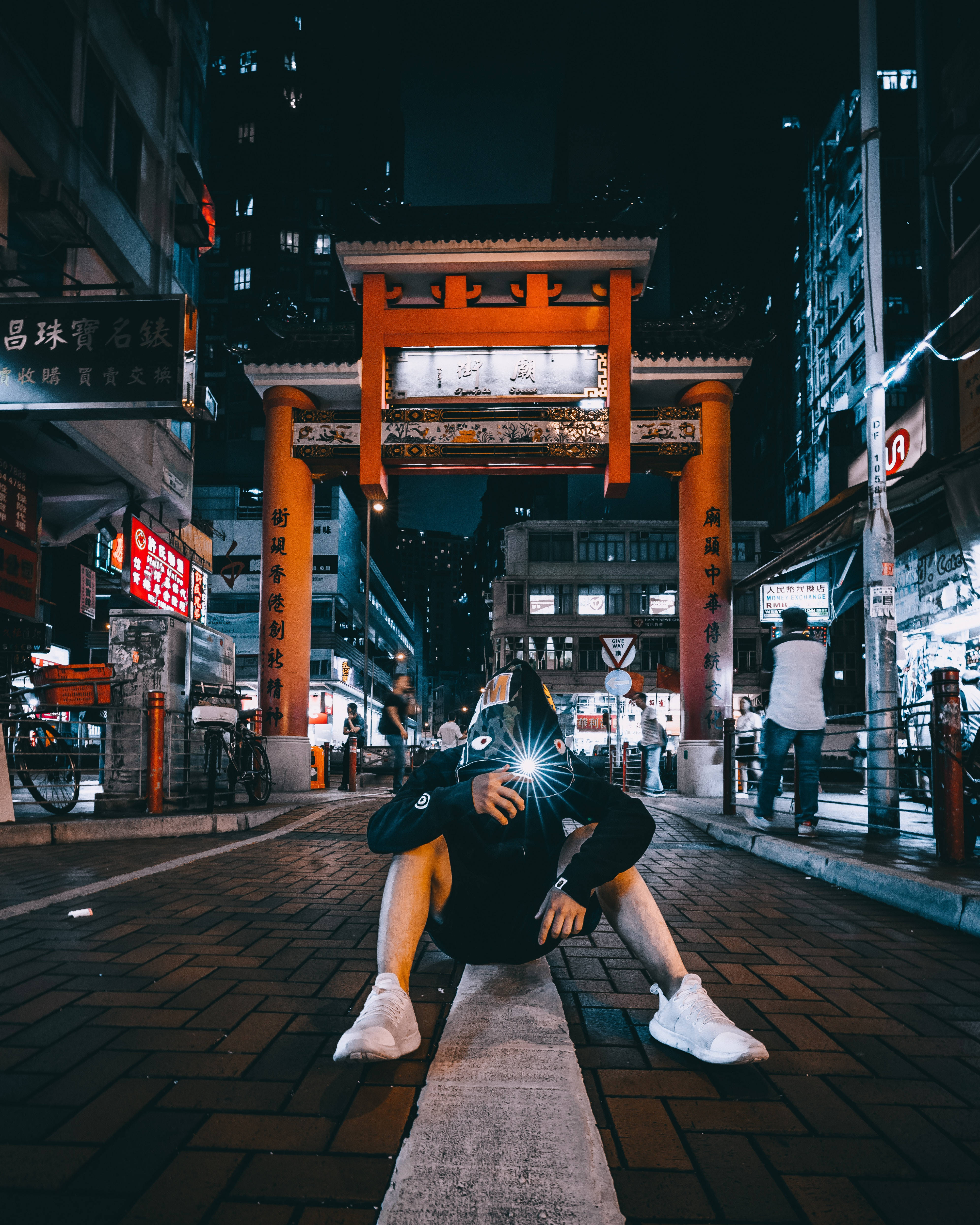 匿名 桌布 壁紙 — 一個人在夜晚蹲在城市街道上，在傳統橙色門下拿著發光手機，周圍有霓虹燈牌發光。