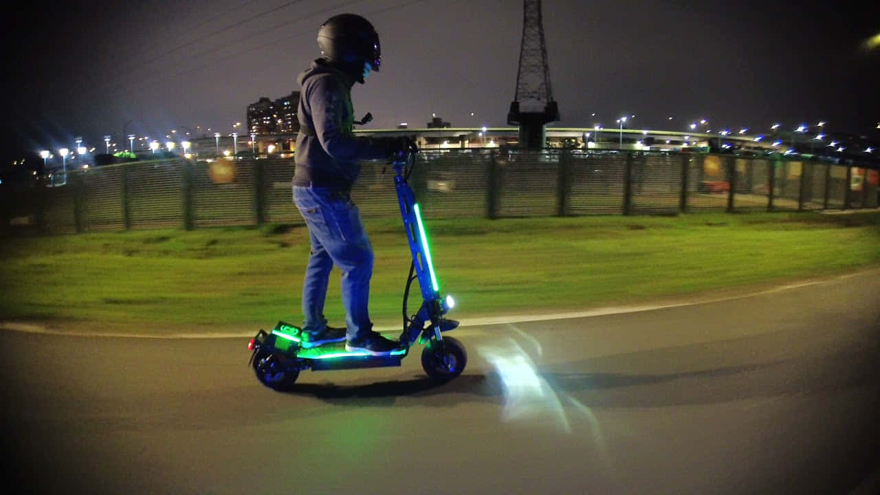 scooter 桌布 壁紙 — 一個人在夜間沿著鋪砌的小路騎著發光的電動滑板車，背景有城市燈光和起重機。