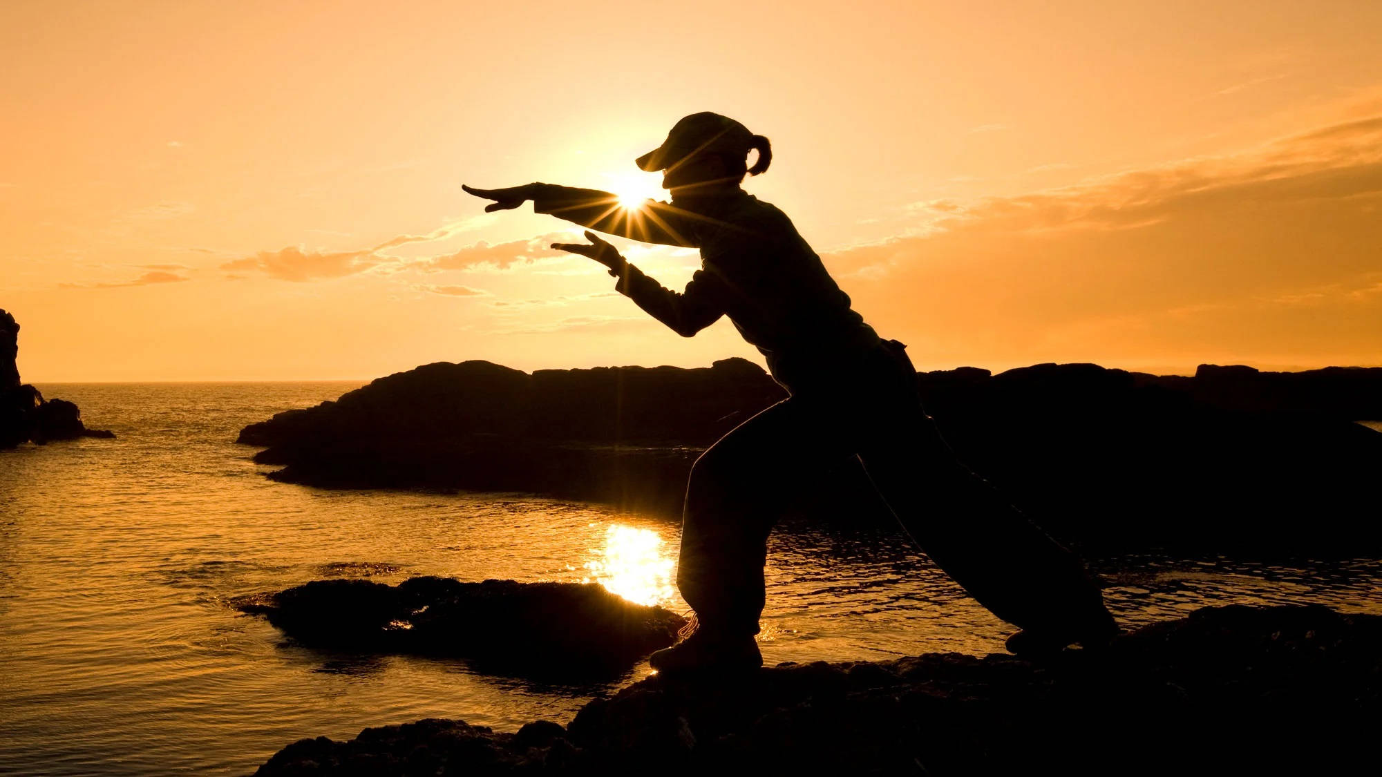Silhouette 桌布 壁紙 — 一個人在岩石海岸上練習武術的剪影，背景是金色日落和太陽爆發的光芒。