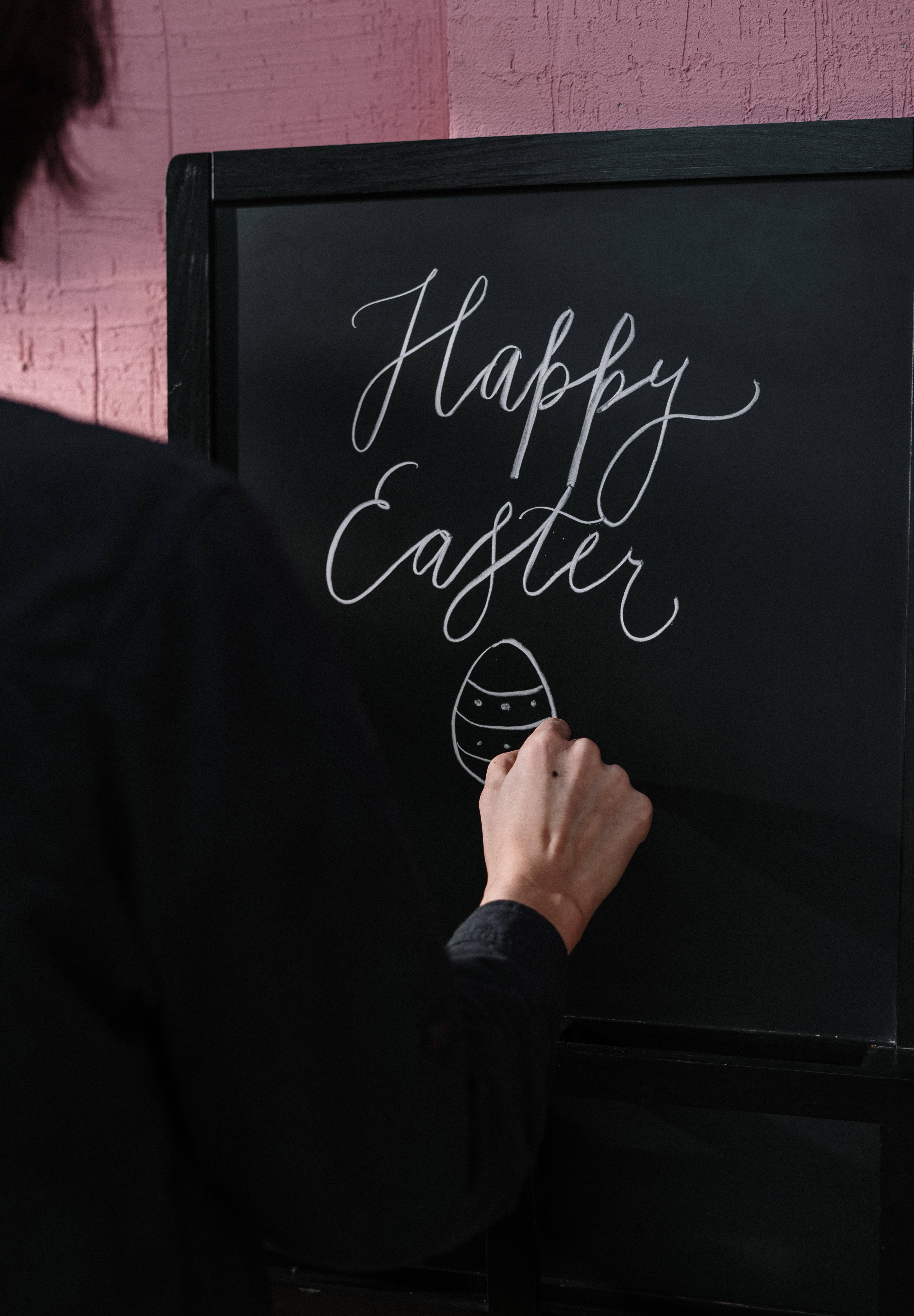 Happy Easter 桌布 壁紙 — 一個人在帶有紋理的粉紅色牆壁前的黑色黑板上用優雅的白色草書寫下「復活節快樂」。