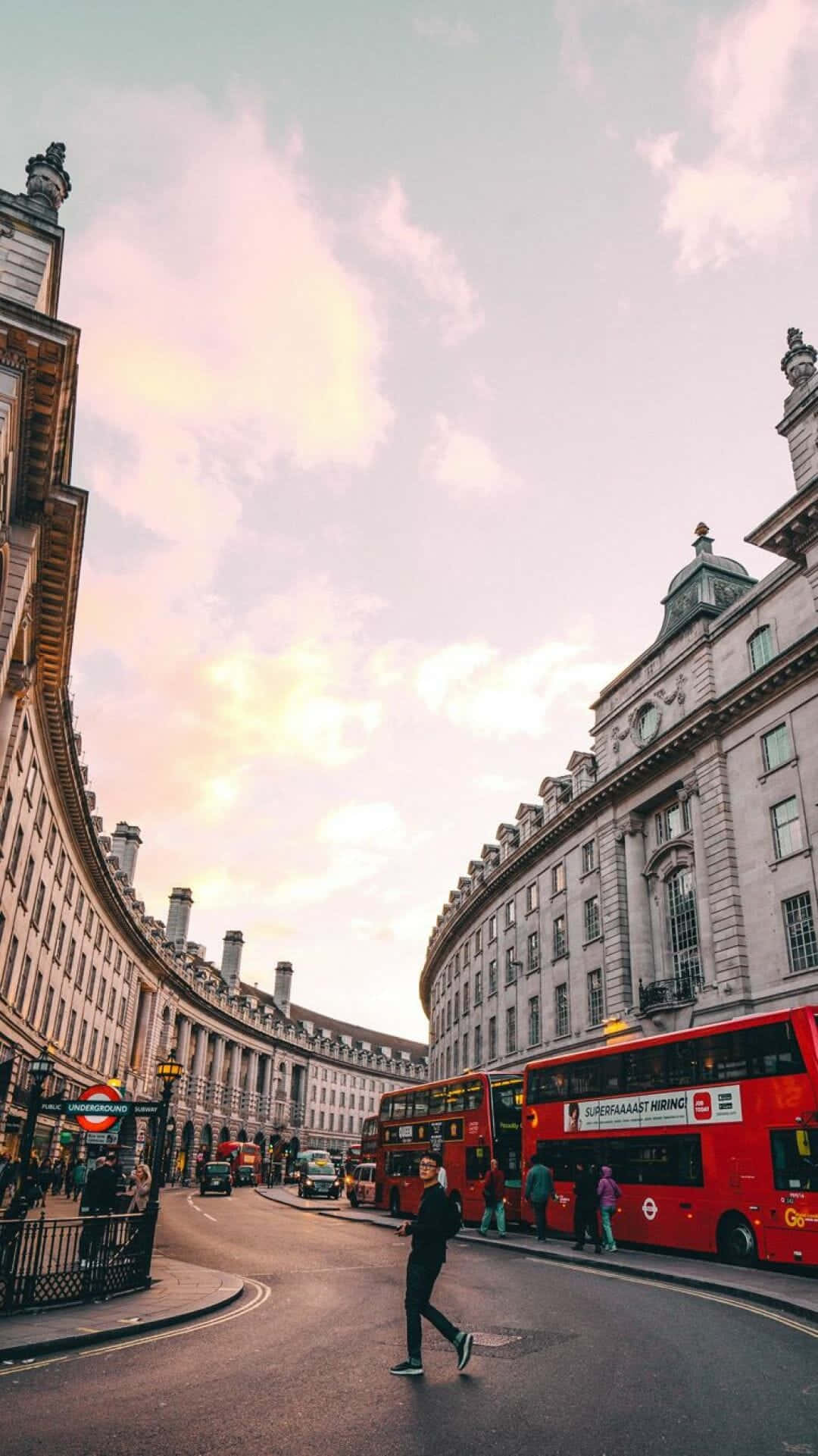 London iPhone 桌布 壁紙 — 一個人在彎曲的倫敦街道上行走，街道兩旁是歷史悠久的石建築和標誌性的紅色雙層巴士，天空呈現柔和的粉紅色和金色日落。