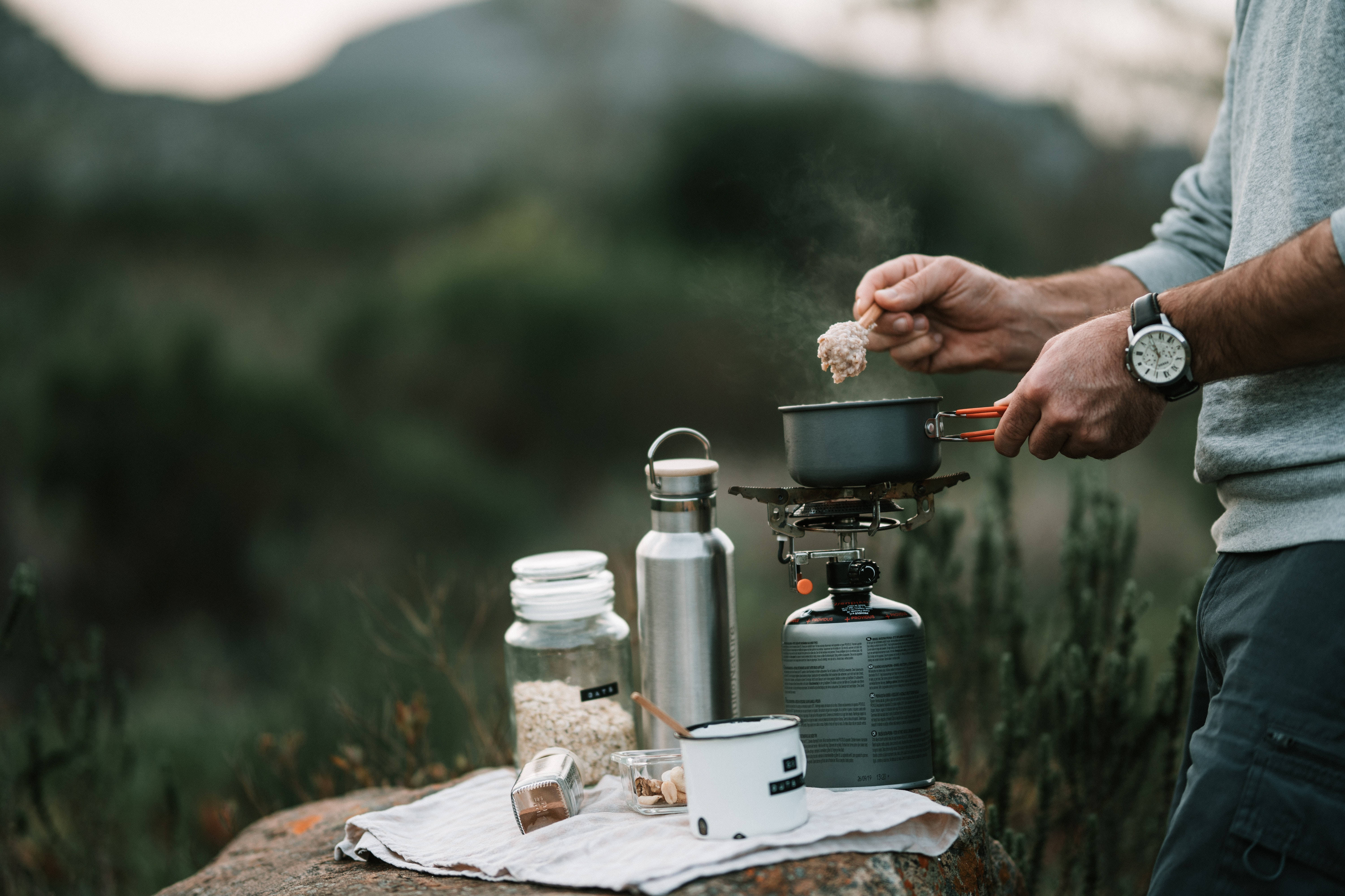 烹飪 桌布 壁紙 — 一個人在戶外使用便攜式爐灶煮食，背景是柔和的山巒。
