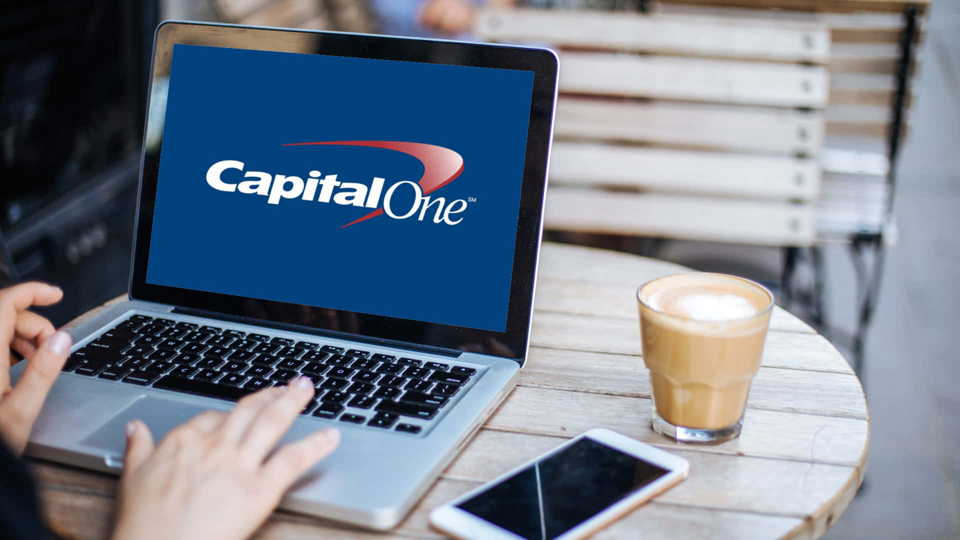 咖啡動圖 桌布 壁紙 — 一個人在戶外咖啡桌旁使用顯示Capital One標誌的筆記型電腦，旁邊有拿鐵和手機。