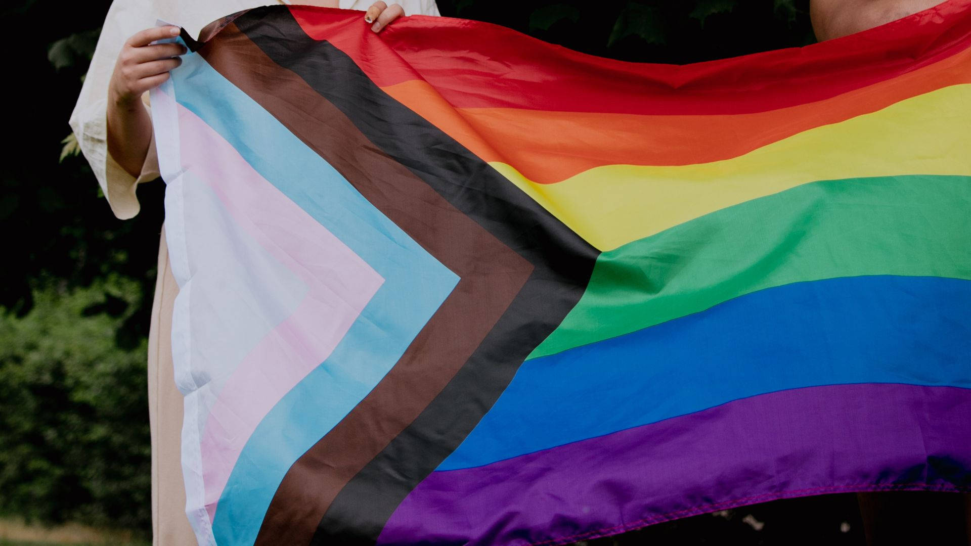 Subtle Lgbt 桌布 壁紙 — 一個人在戶外柔和自然光下，手持鮮豔的彩虹和棕色跨性別旗幟，周圍綠意盎然。