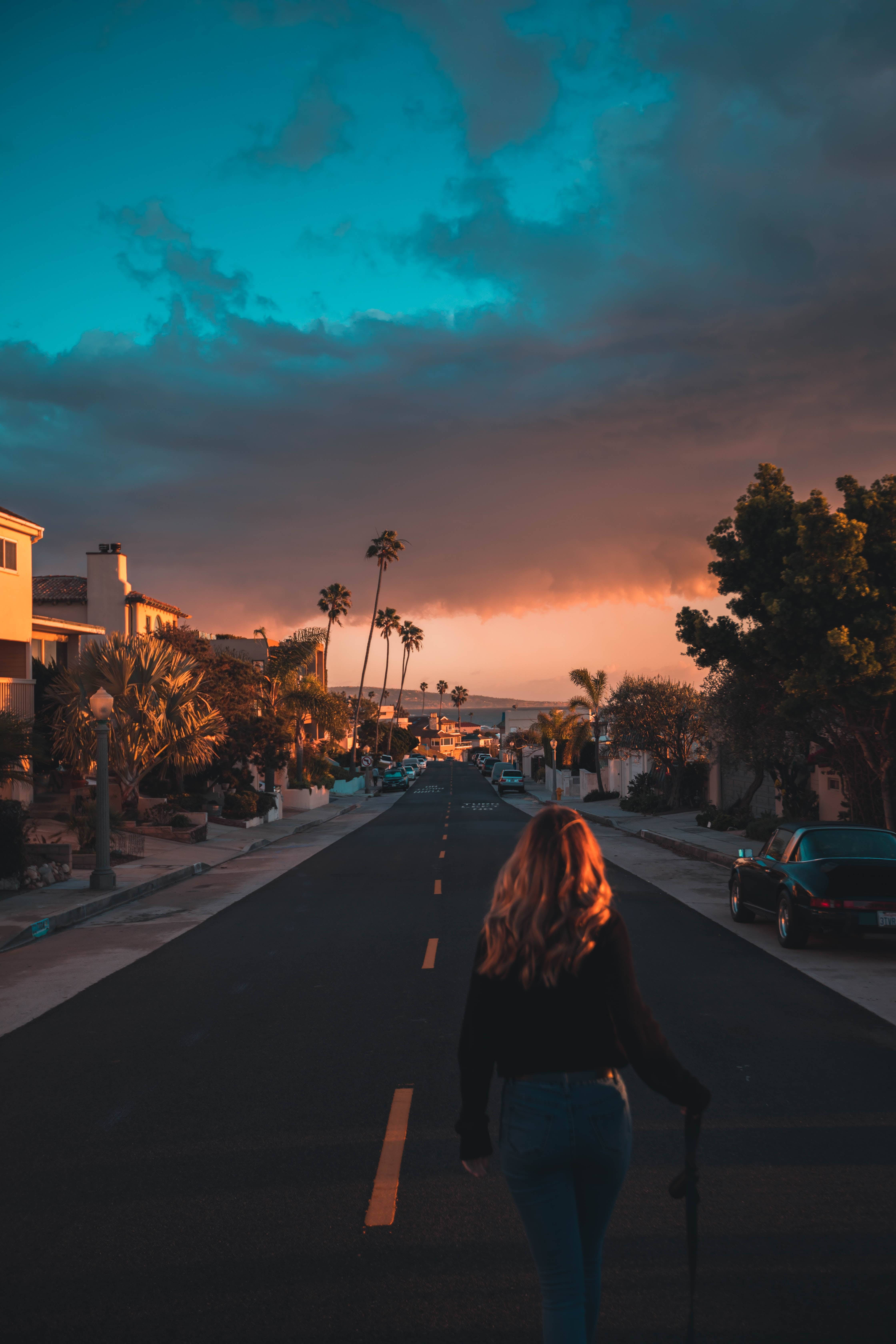 Boulevard 桌布 壁紙 — 一個人在日落時分沿著一條寧靜的街道行走，棕櫚樹和房屋在戲劇性的藍色和橙色天空下。