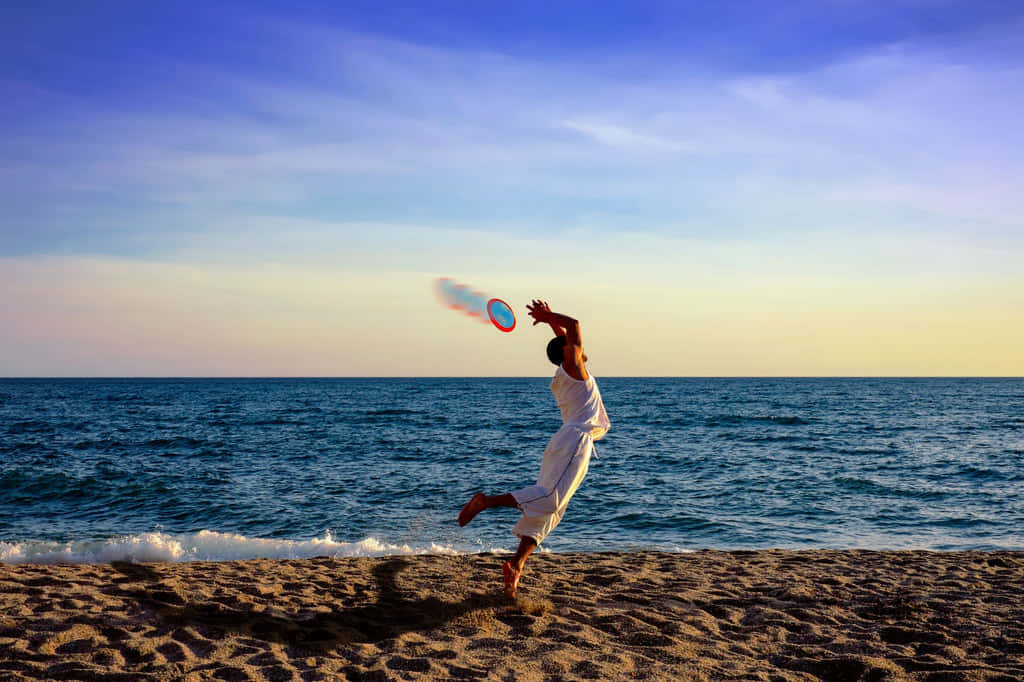 Beach Aesthetic 桌布 壁紙 — 一個人在日落時分的沙灘上快樂地跳躍，在充滿活力的藍色和橙色天空下接住飛盤。