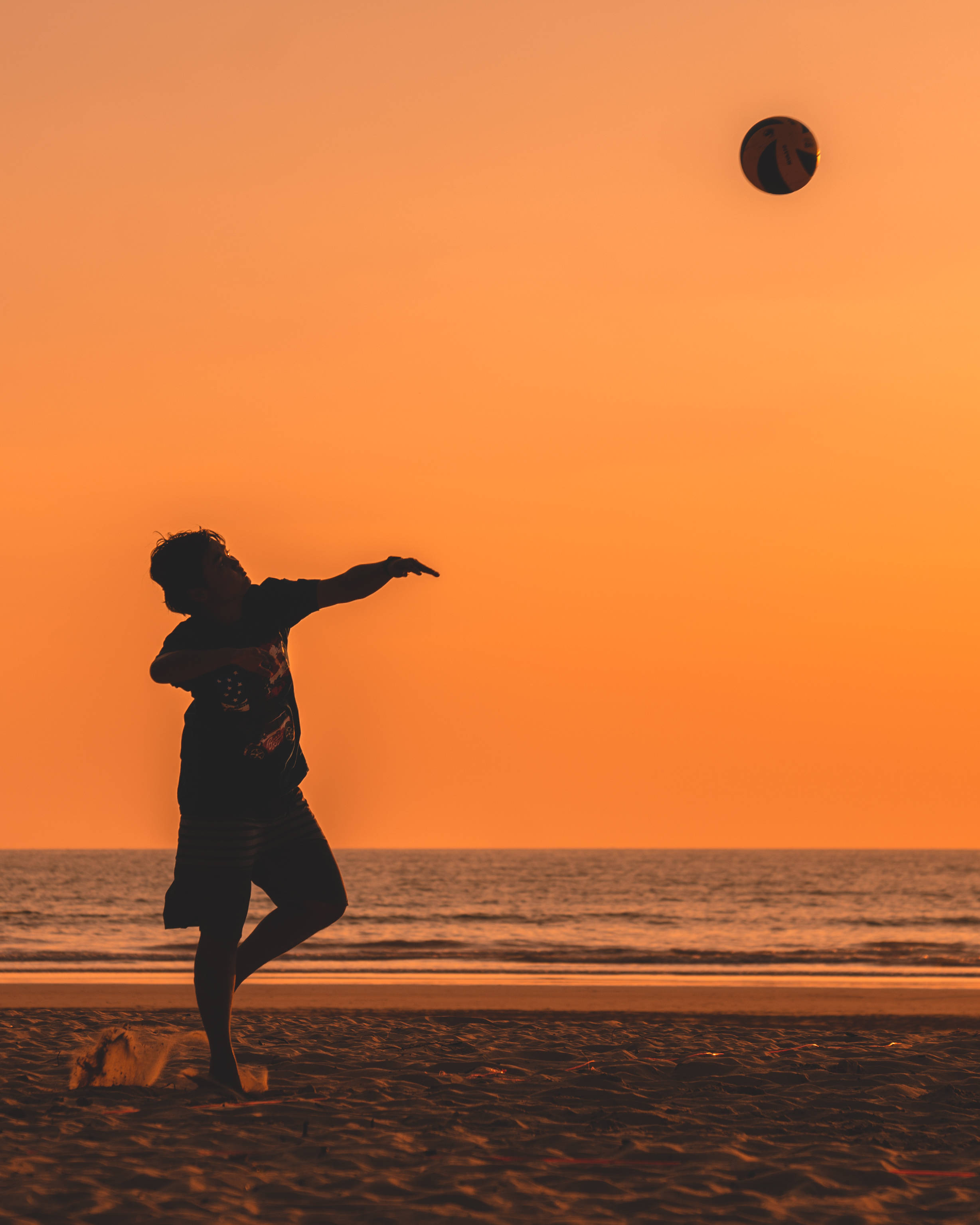 volleyball 4K 桌布 壁紙 — 一個人在日落時分的沙灘上踢足球的剪影，溫暖的橙色天空和海洋波浪。