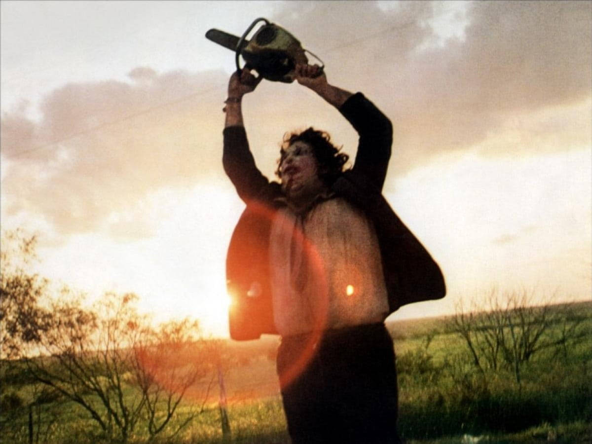 Leatherface 桌布 壁紙 — 一個人在日落時分的田野中，高舉電鋸，映襯著發光的天空。