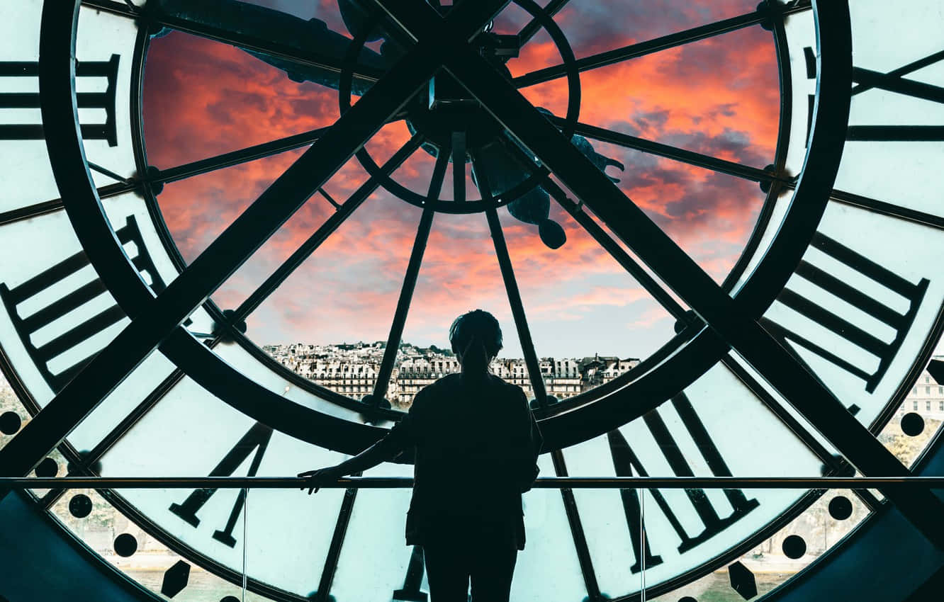 Silhouette 桌布 壁紙 — 一個人在日落時分站在巨大鐘面前的剪影，被羅馬數字框住，背景是火紅的橙色天空。