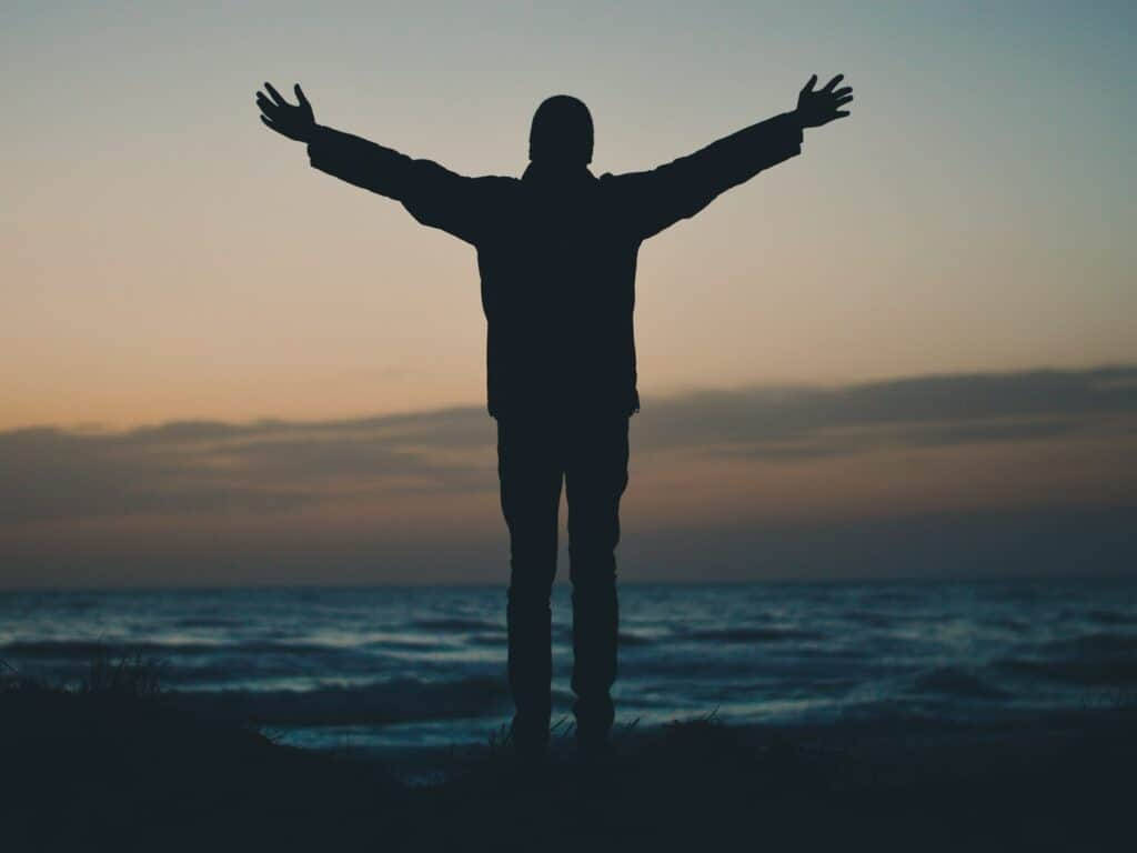 Man 桌布 壁紙 — 一個人在日落時分站在沙灘上張開雙臂，在柔和的橙色和藍色天空下形成一個深色剪影。