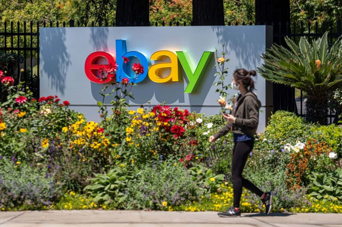 eBay 桌布 壁紙 — 一個人在明亮的日光下走過一個充滿活力的eBay標誌，周圍環繞著色彩繽紛的花朵和綠植。