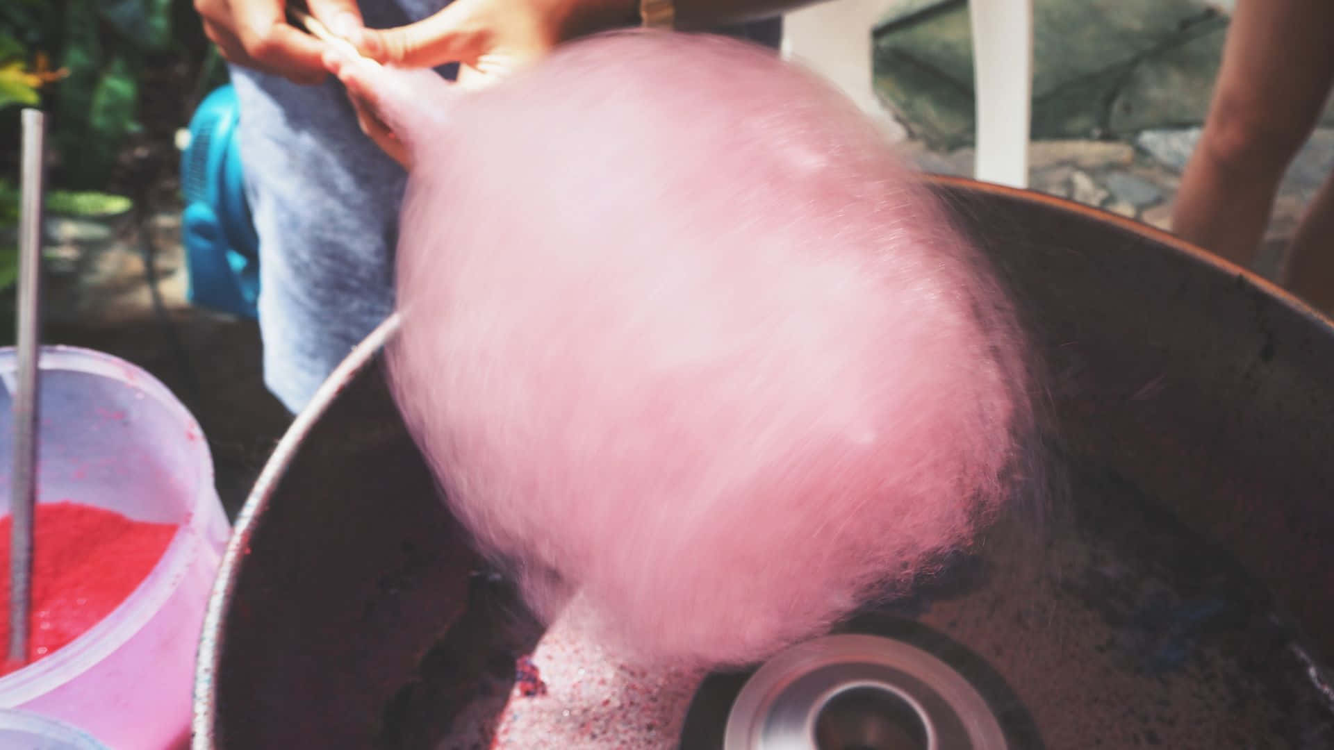 Cotton Candy 桌布 壁紙 — 一個人在明亮的陽光下，從戶外旋轉的機器上拉出 fluffy 粉紅色棉花糖。