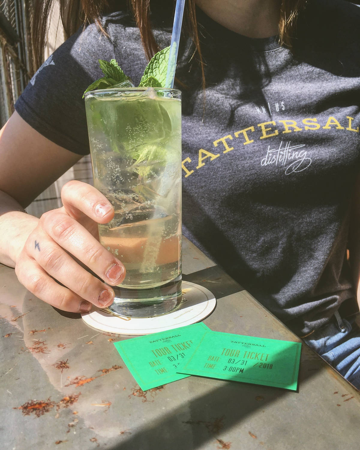 Drink 桌布 壁紙 — 一個人在明媚陽光下手持薄荷雞尾酒，綠色旅遊門票放在一張風化的桌子上。