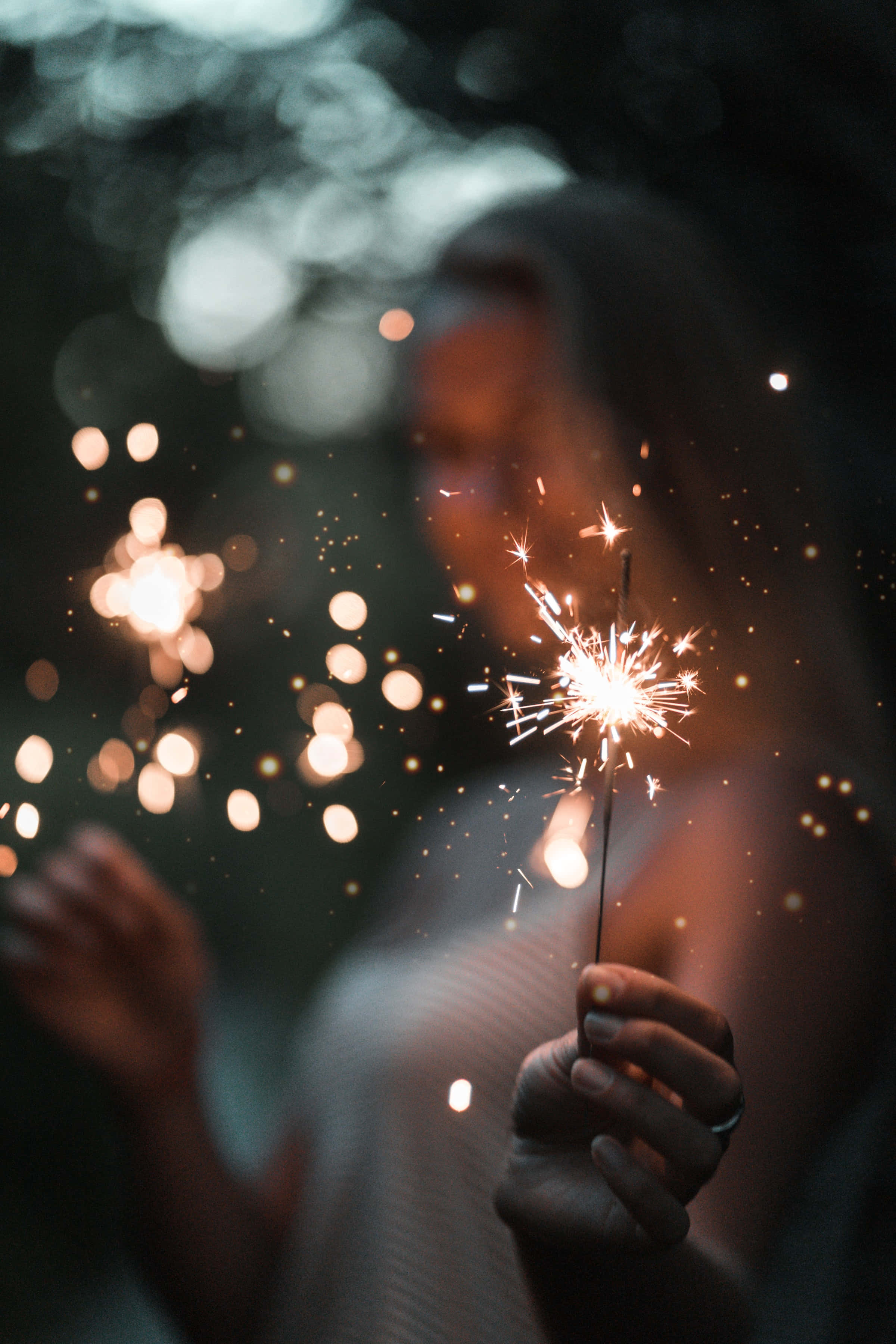 sparklers 桌布 壁紙 — 一個人在昏暗模糊的背景中，手持閃爍的煙火，背景有散景燈光。
