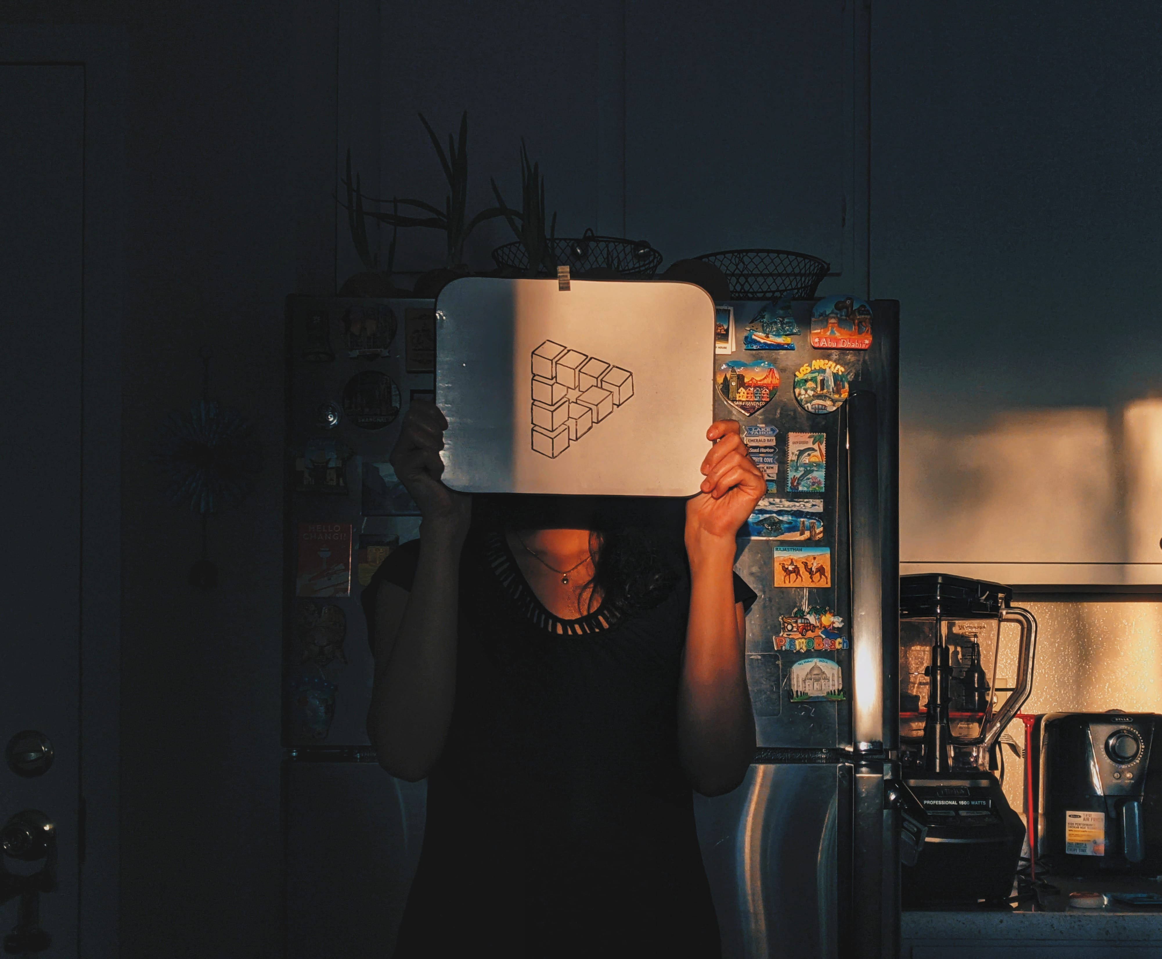 Illusion 桌布 壁紙 — 一個人在昏暗的廚房裡拿著一個金屬托盤，上面有3D立方體錯覺遮住臉，在佈滿磁鐵的冰箱上投下戲劇性的陰影。