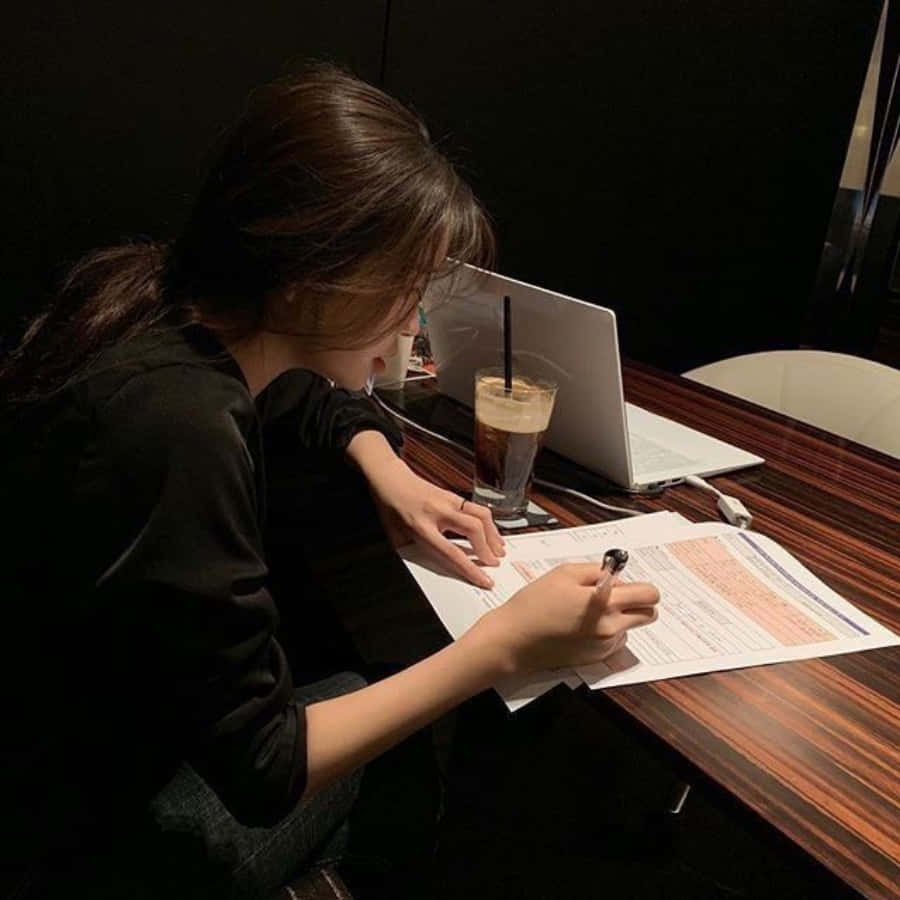 Study Girl 桌布 壁紙 — 一個人在昏暗的房間裡，俯身在一張木桌上，在筆記型電腦和冰咖啡旁邊寫字。