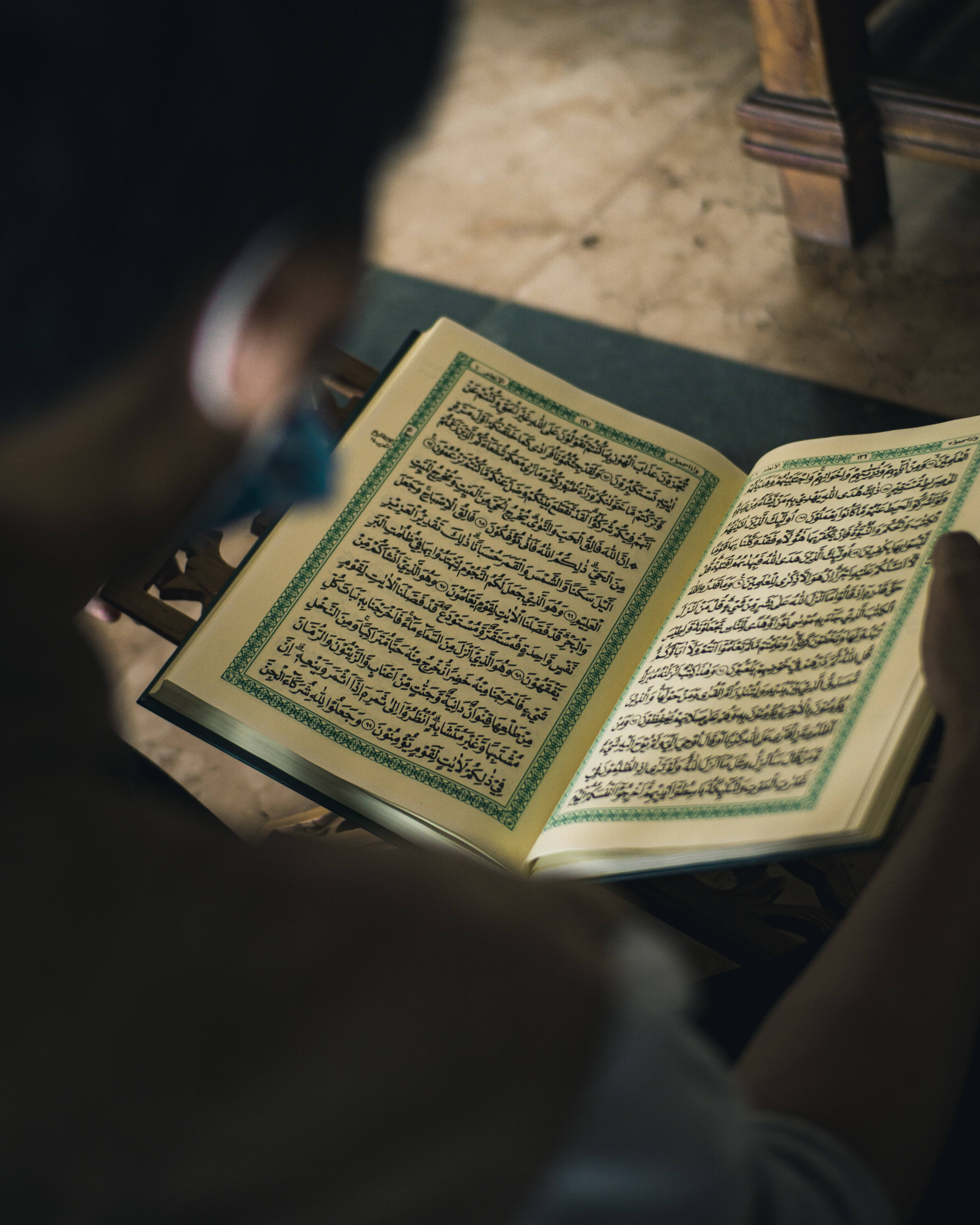 Best Islamic 桌布 壁紙 — 一個人在昏暗的房間裡，在溫暖的燈光下拿著一本打開的、頁面邊緣為綠色的古蘭經。