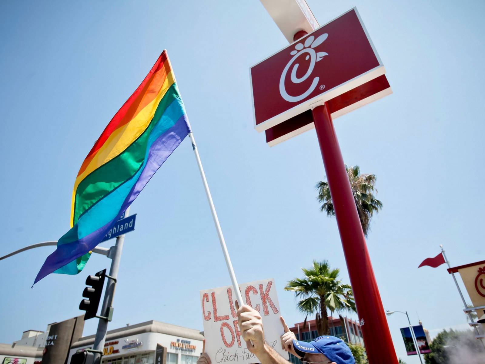 chick 桌布 壁紙 — 一個人在晴朗的藍天下，在Chick-fil-A餐廳前舉著彩虹旗和抗議標語。