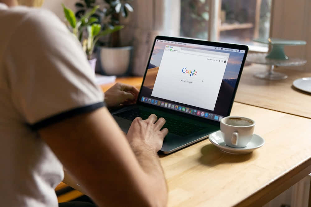 google 桌布 壁紙 — 一個人在木桌上打字，旁邊有咖啡杯和盆栽，柔和自然光線。