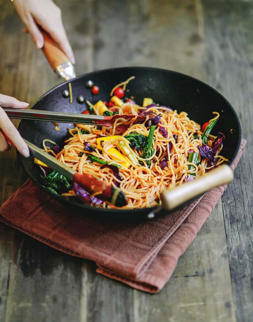Food Photography 桌布 壁紙 — 一個人在木桌上的鏽色毛巾上的黑色鍋中攪拌色彩繽紛的蔬菜麵條。