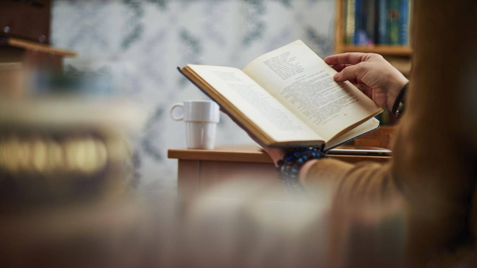 書本美學 桌布 壁紙 — 一個人在木桌上讀書，旁邊有一個白色杯子，在舒適的室內環境中。