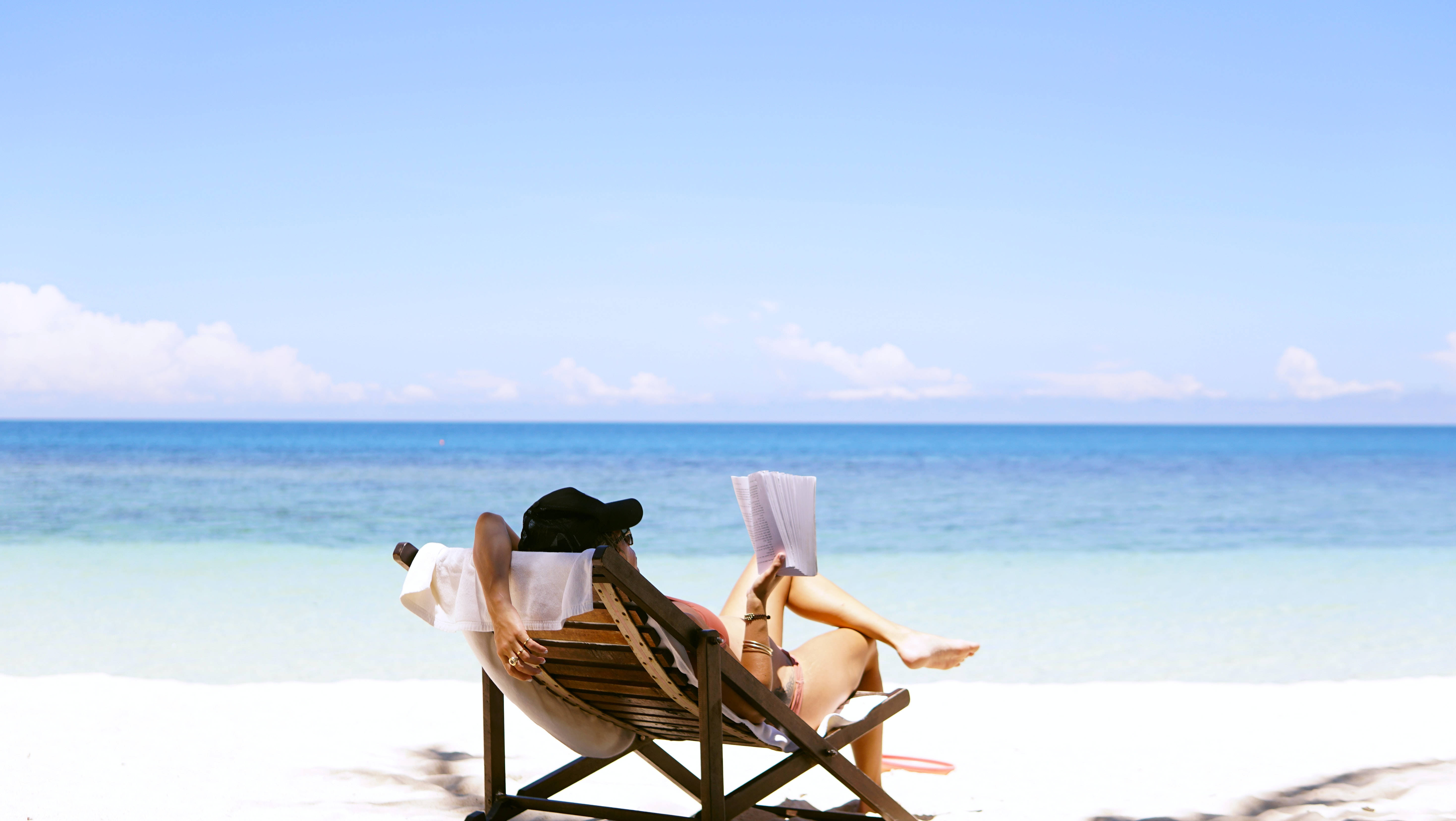 Beach Scene 桌布 壁紙 — 一個人在木製沙灘椅上放鬆，在清澈的藍天下閱讀一本書，綠松石色的海洋一直延伸到地平線。