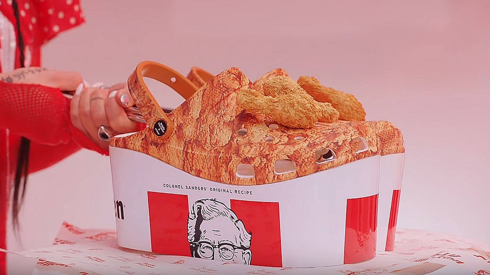 Crocs 桌布 壁紙 — 一個人在柔和粉紅色背景前，拿著一個裝滿炸雞的KFC桶形木屐。