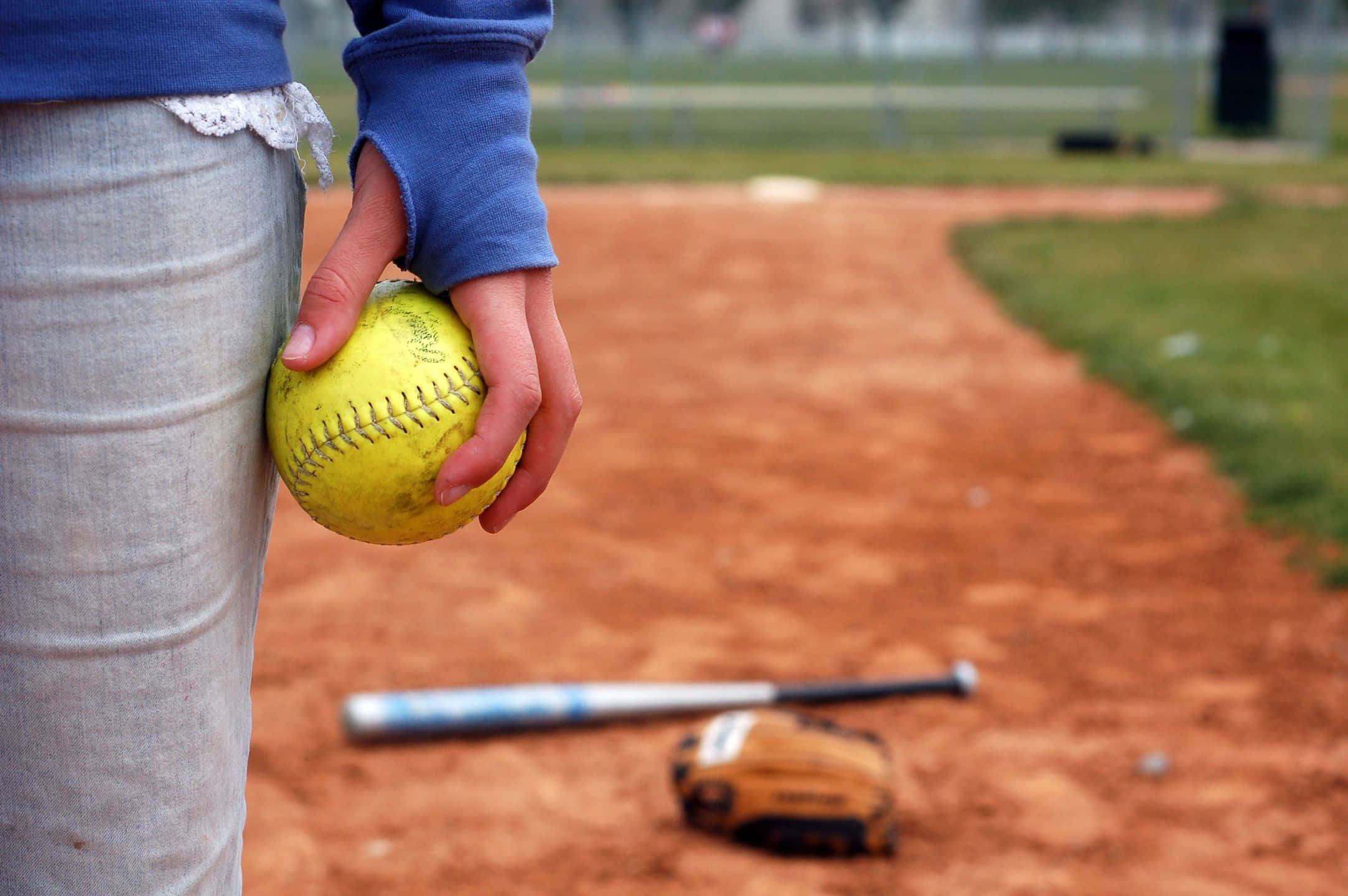 Little League Baseball 桌布 壁紙 — 一個人在棒球場上手持黃色壘球的特寫，球棒和手套放在泥土上。