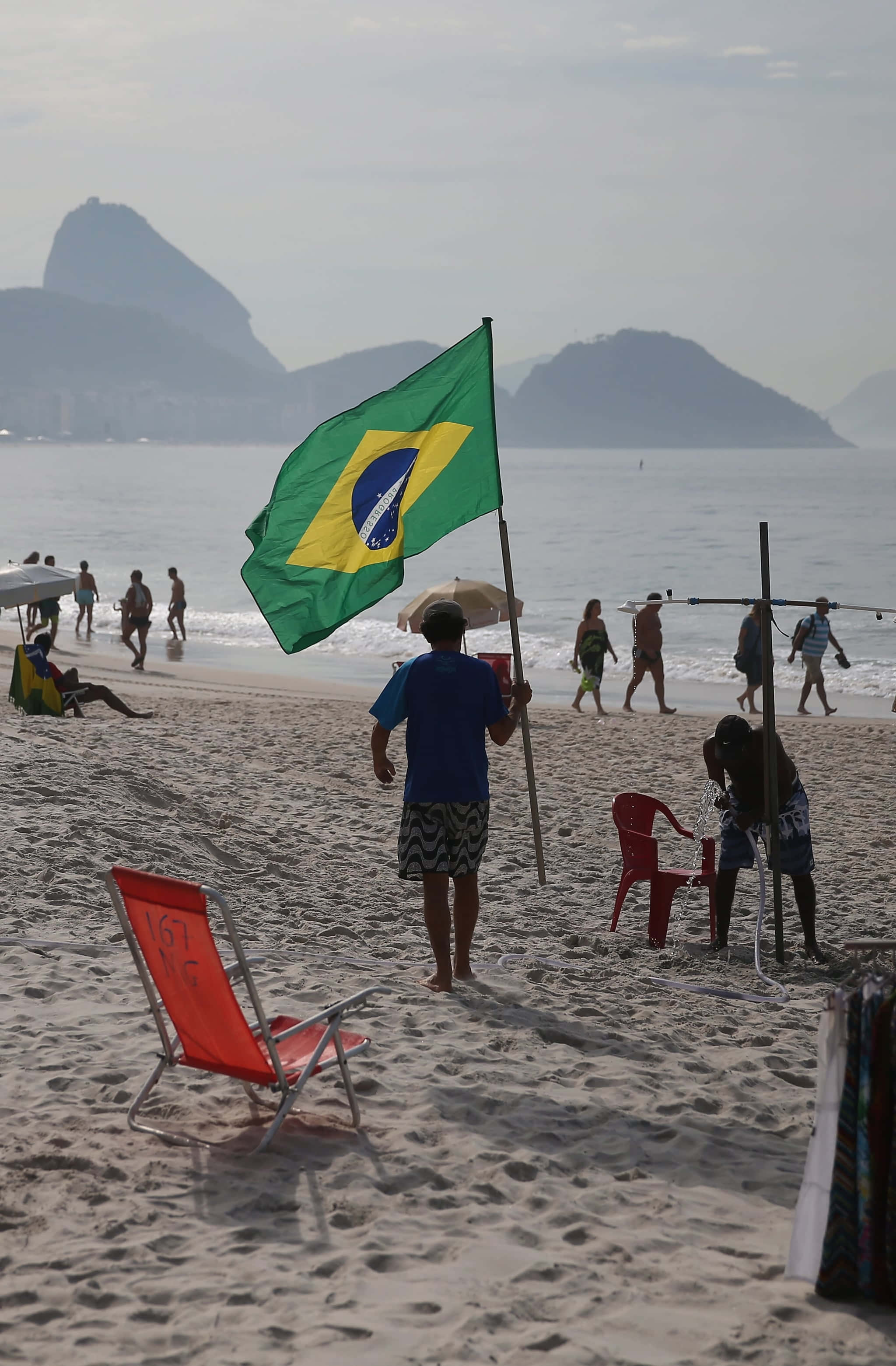 Brazilian Beach 桌布 壁紙 — 一個人在沙灘上手持巴西國旗，遠方糖麵包山在薄霧中隱約可見，海灘遊客在岸邊漫步。