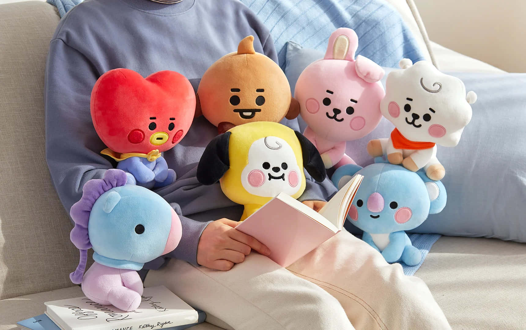 BT21 桌布 壁紙 — 一個人在沙發上放鬆，周圍擺滿色彩繽紛的BT21絨毛玩具，同時閱讀一本書。