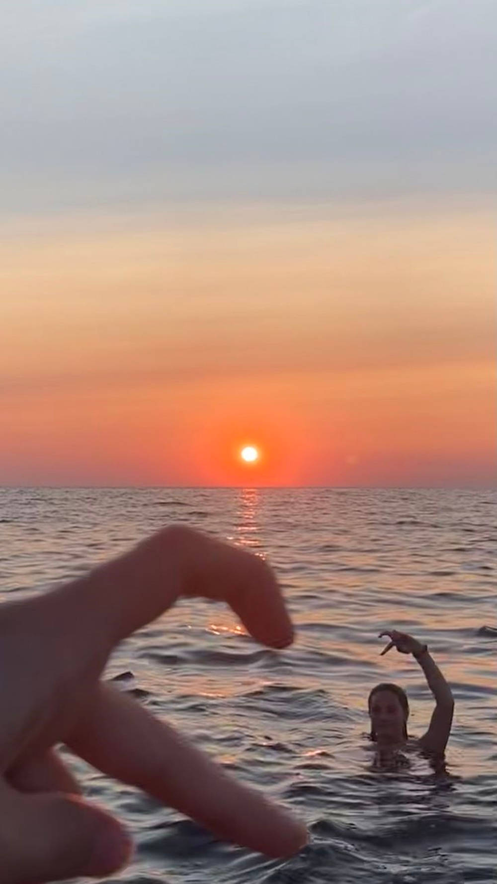 日落 桌布 壁紙 — 一個人在海洋中游泳，一隻手框住場景，背景是鮮豔的橙色日落。